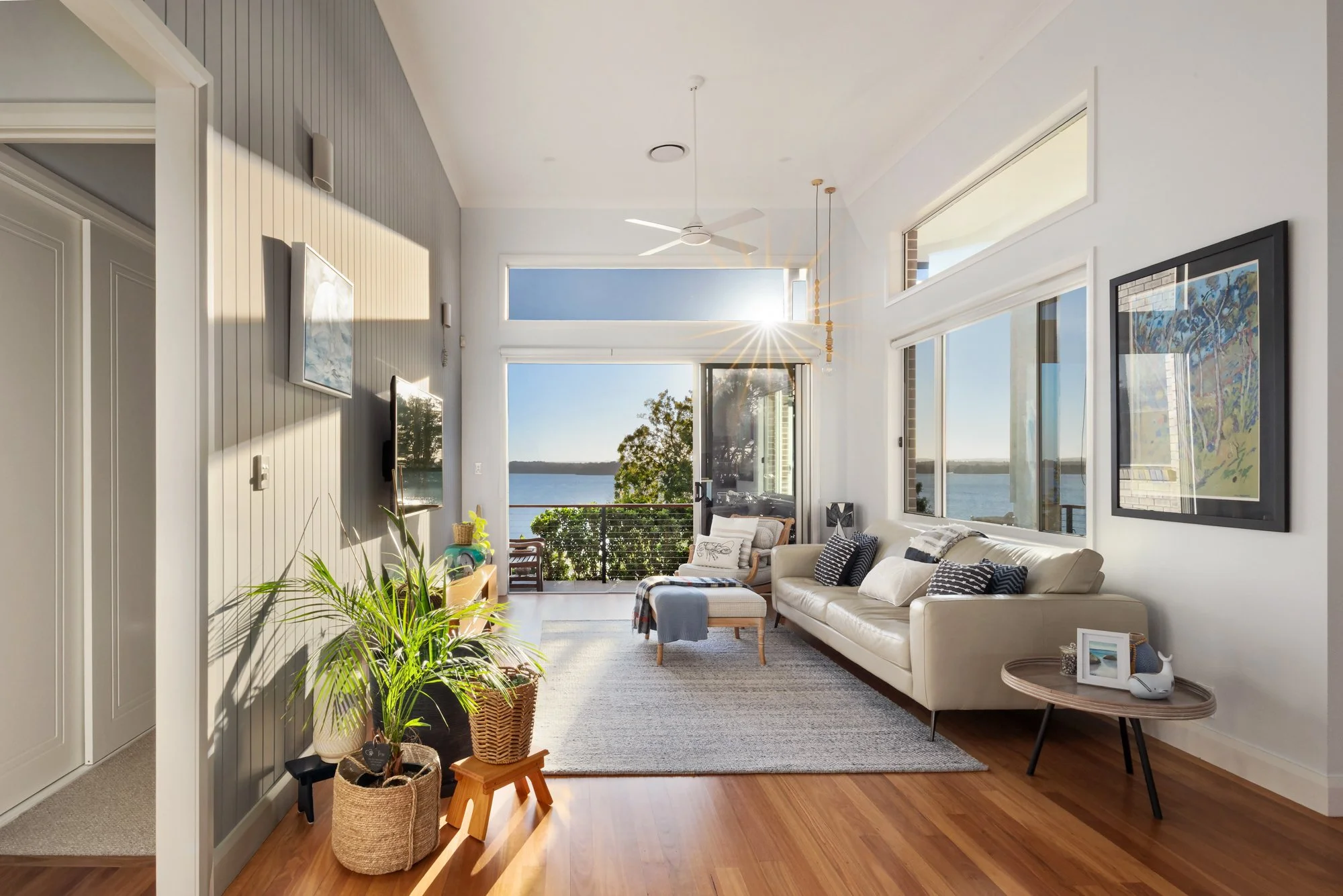 Bright living room with large windows overlooking water, white couch with pillows, armchair, potted plants, and artwork, with sunlight streaming in.