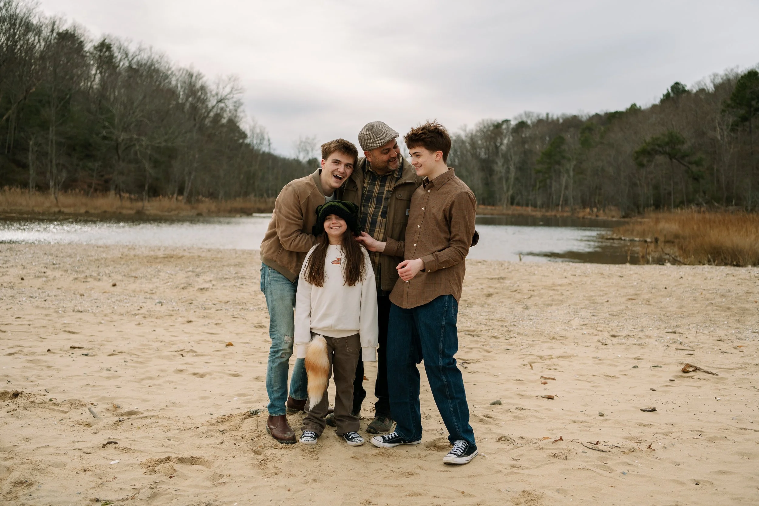 Family laughing together during an outdoor adventure family portrait session at Calvert Cliffs State Park in Southern Maryland, standing on the sandy shoreline with water and trees in the background.