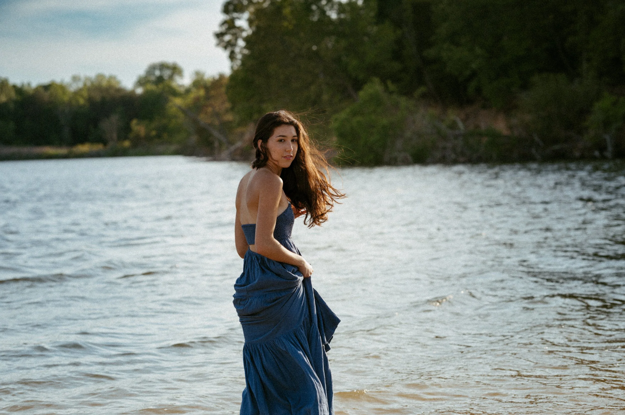 A young woman in a blue dress standing in the water near a shoreline with trees and a cloudy sky in the background.