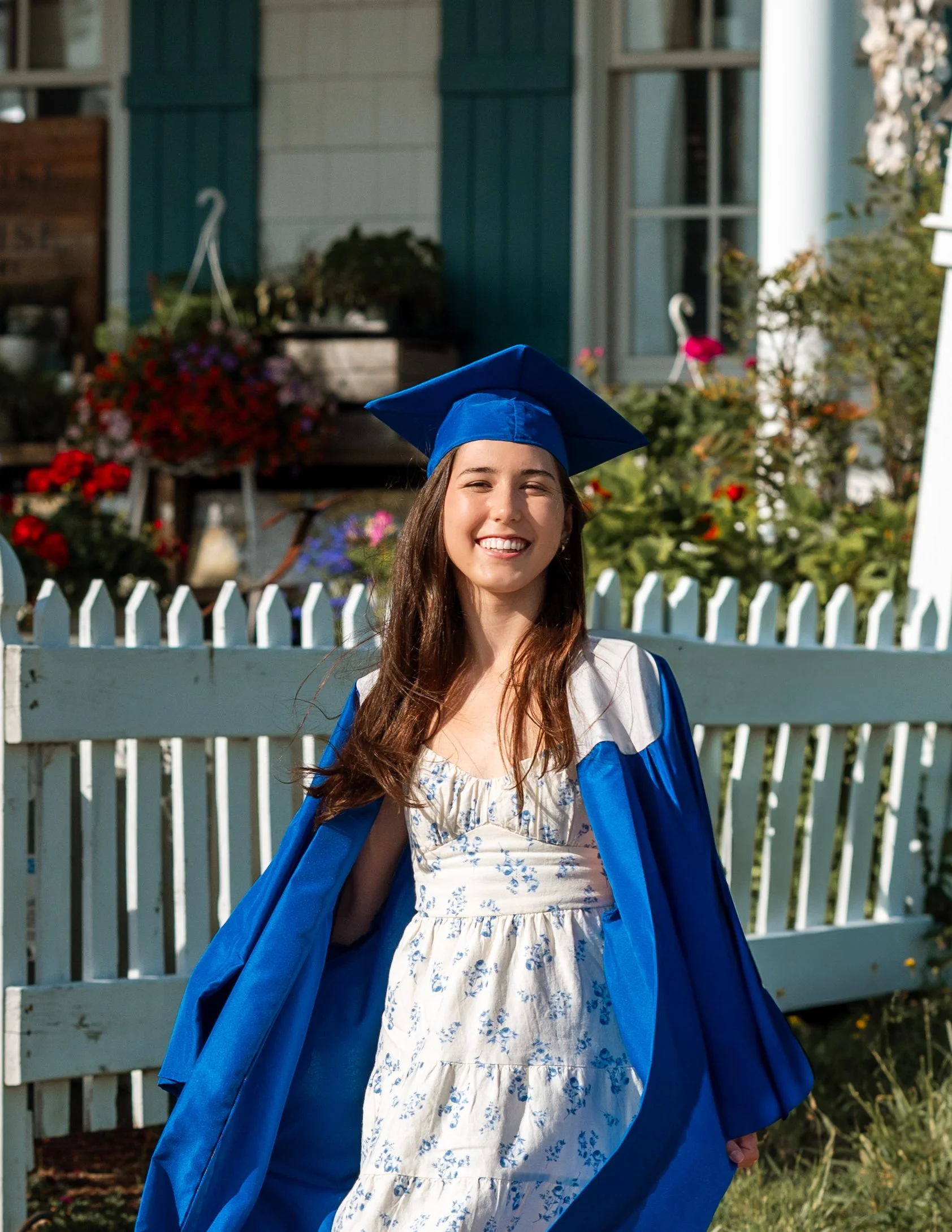 Young woman in graduation cap and gown smiling outdoors in front of a white picket fence and a house with flowers.