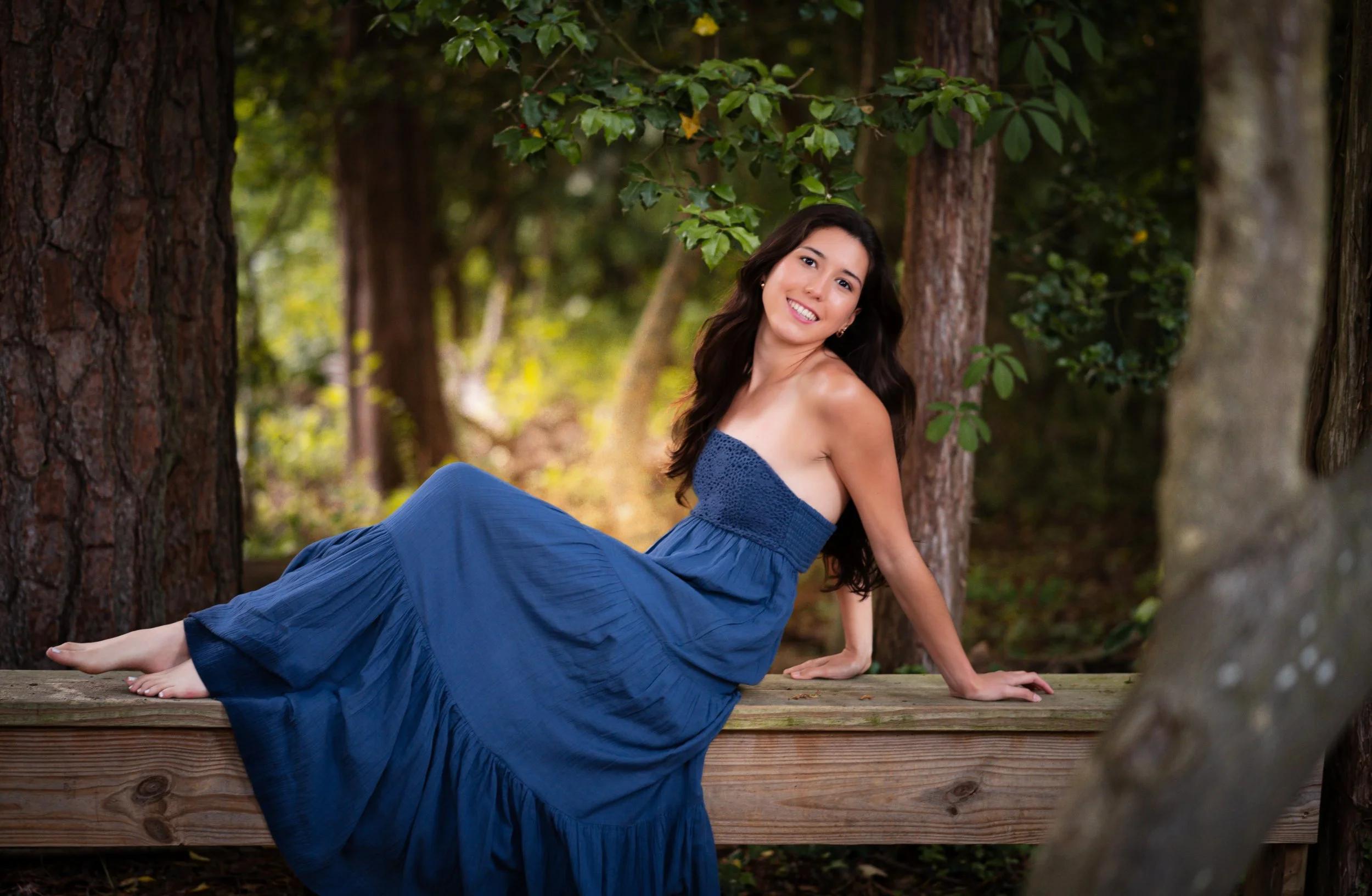 A woman in a strapless blue dress sitting on a wooden platform in a forest.