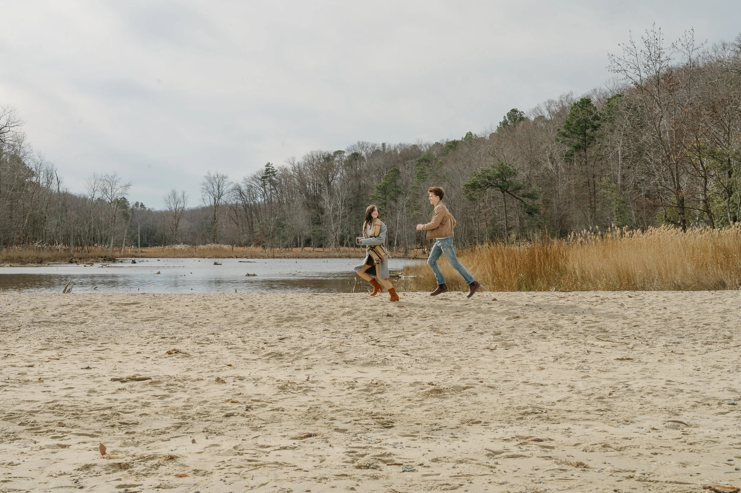Michael and Isabella run along the sandy shoreline at Calvert Cliffs State Park during a winter adventure portrait session, with water, tall grasses, and bare trees in the background.