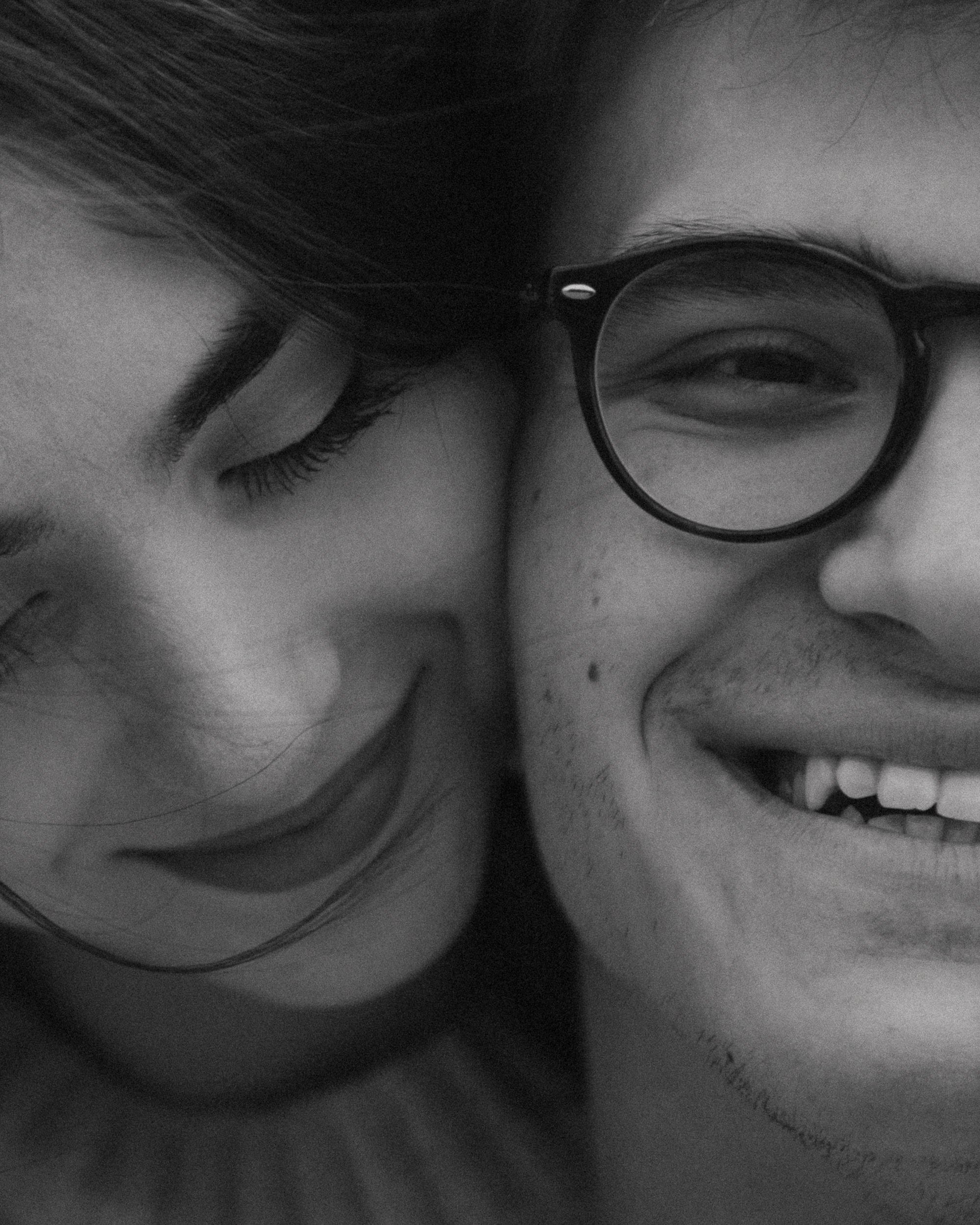 A black and white close-up photo of a smiling couple, with one person wearing glasses and the other with eyes closed, both in a happy moment.