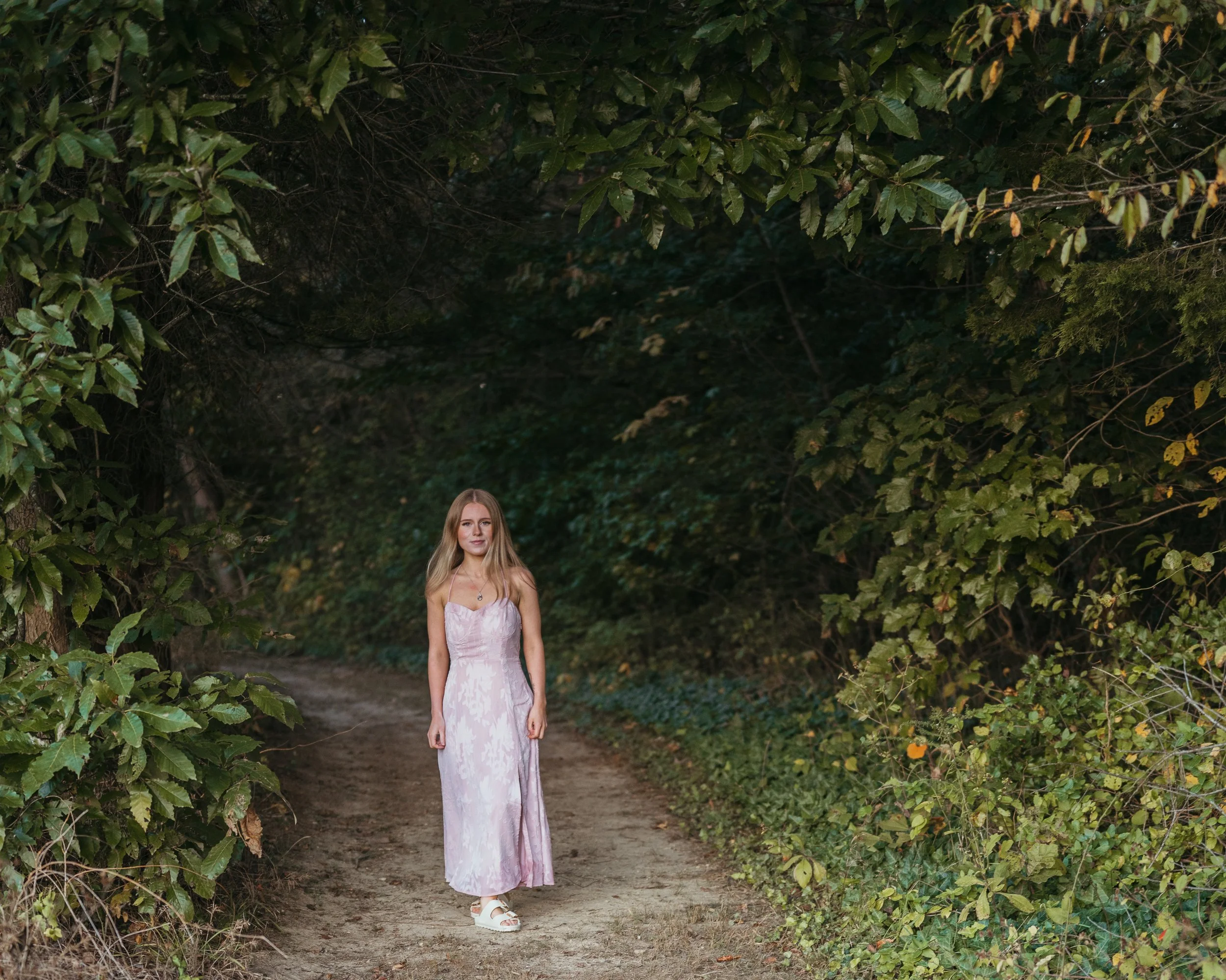 A woman in a pink dress standing on a forest path surrounded by green foliage.