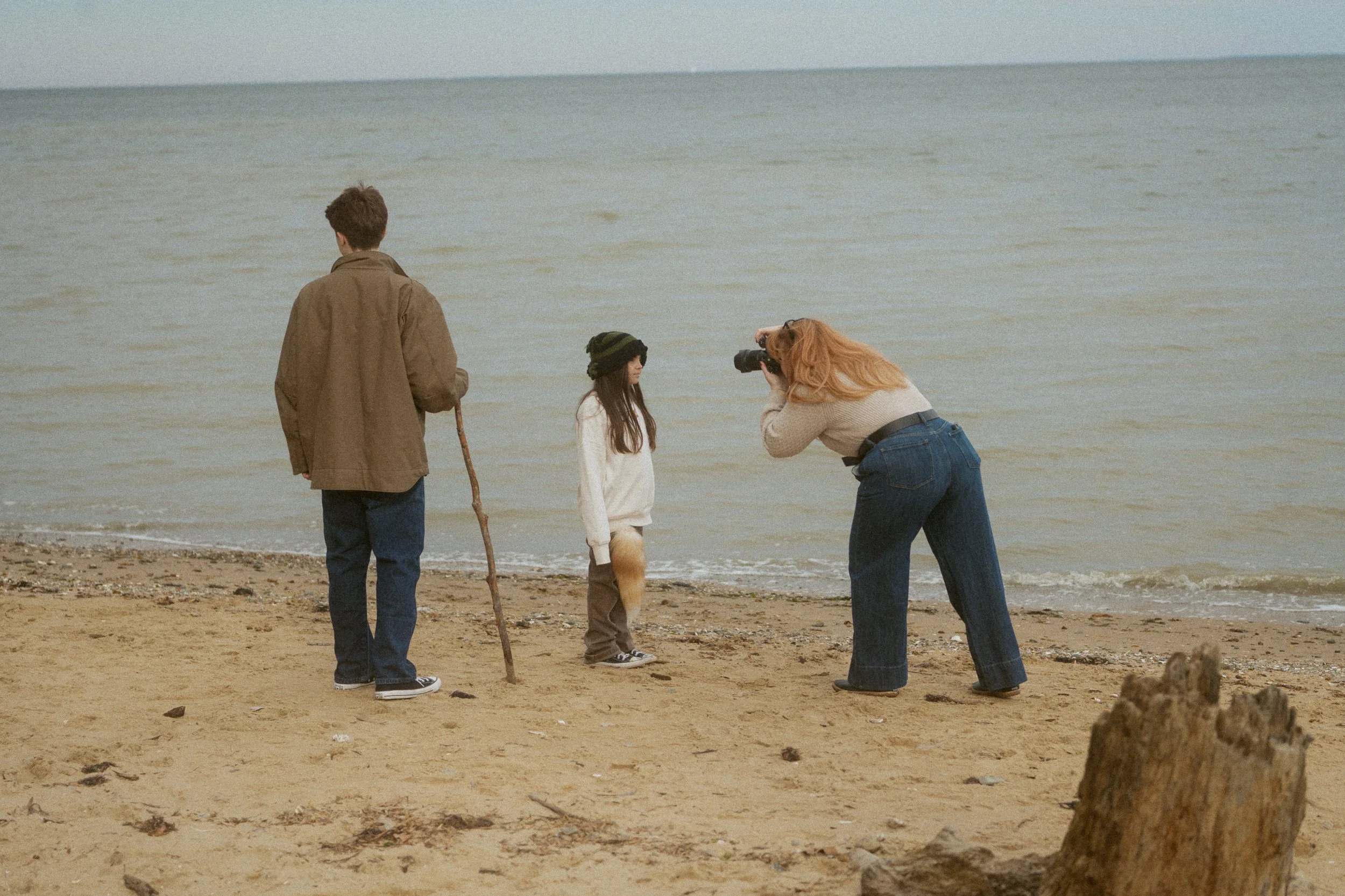 A woman taking a photograph of a girl with a fox tail and a man with a walking stick on the beach.