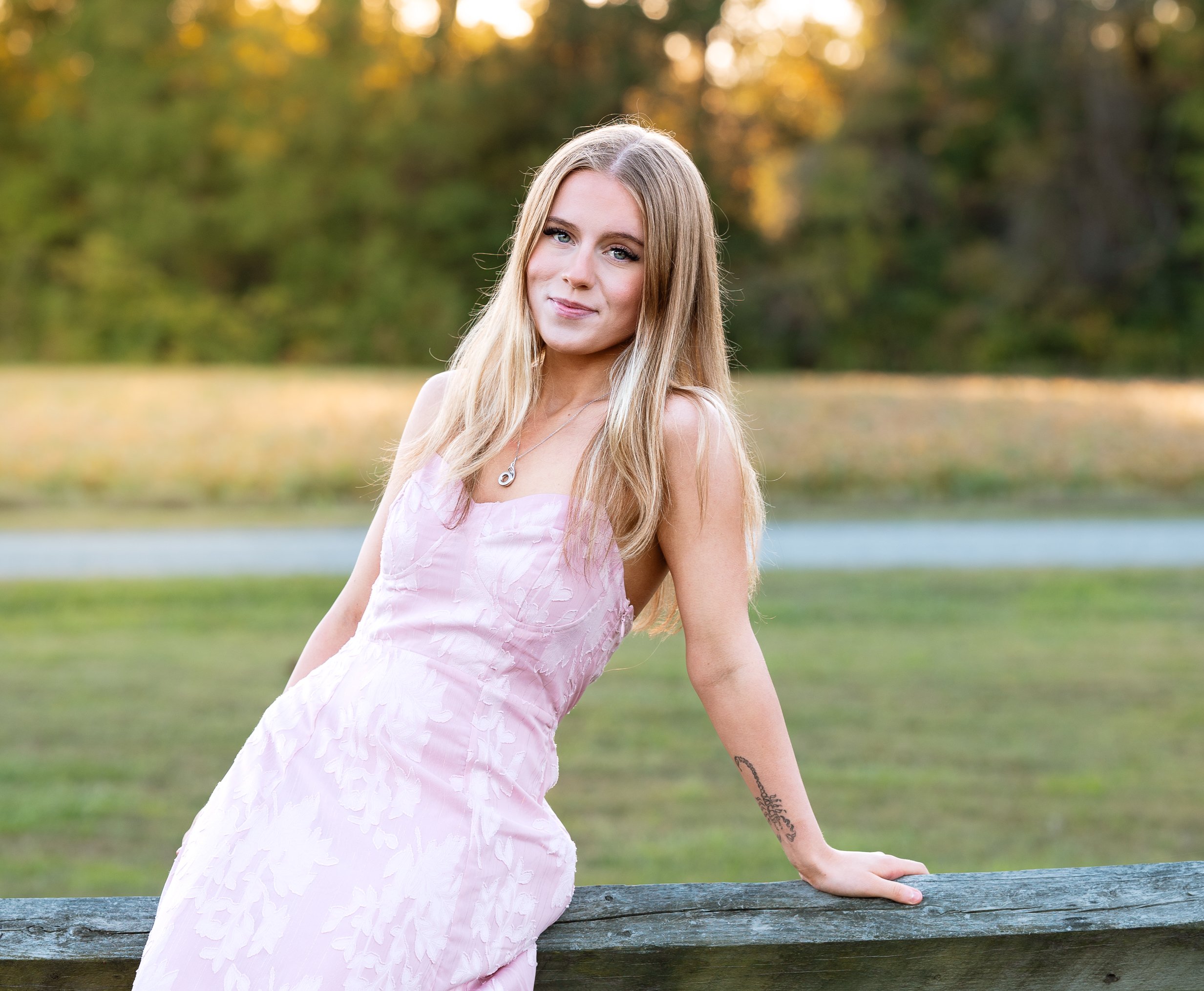 Young woman with long blonde hair in a pink dress stands outdoors leaning on a wooden railing, with trees and a grassy park in the background, during autumn.