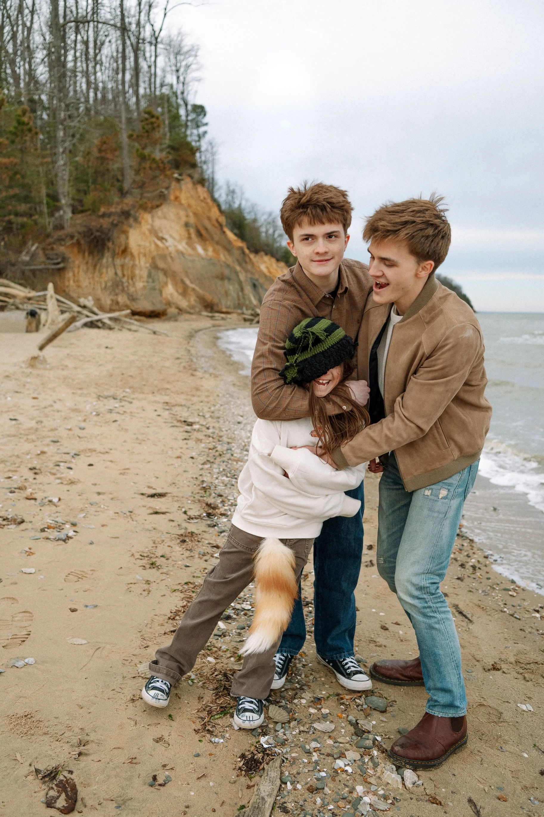 Brothers play with their little sister during a family portrait session at Calvert Cliffs State park as they stand near the shore with the cliffs in the foreground.
