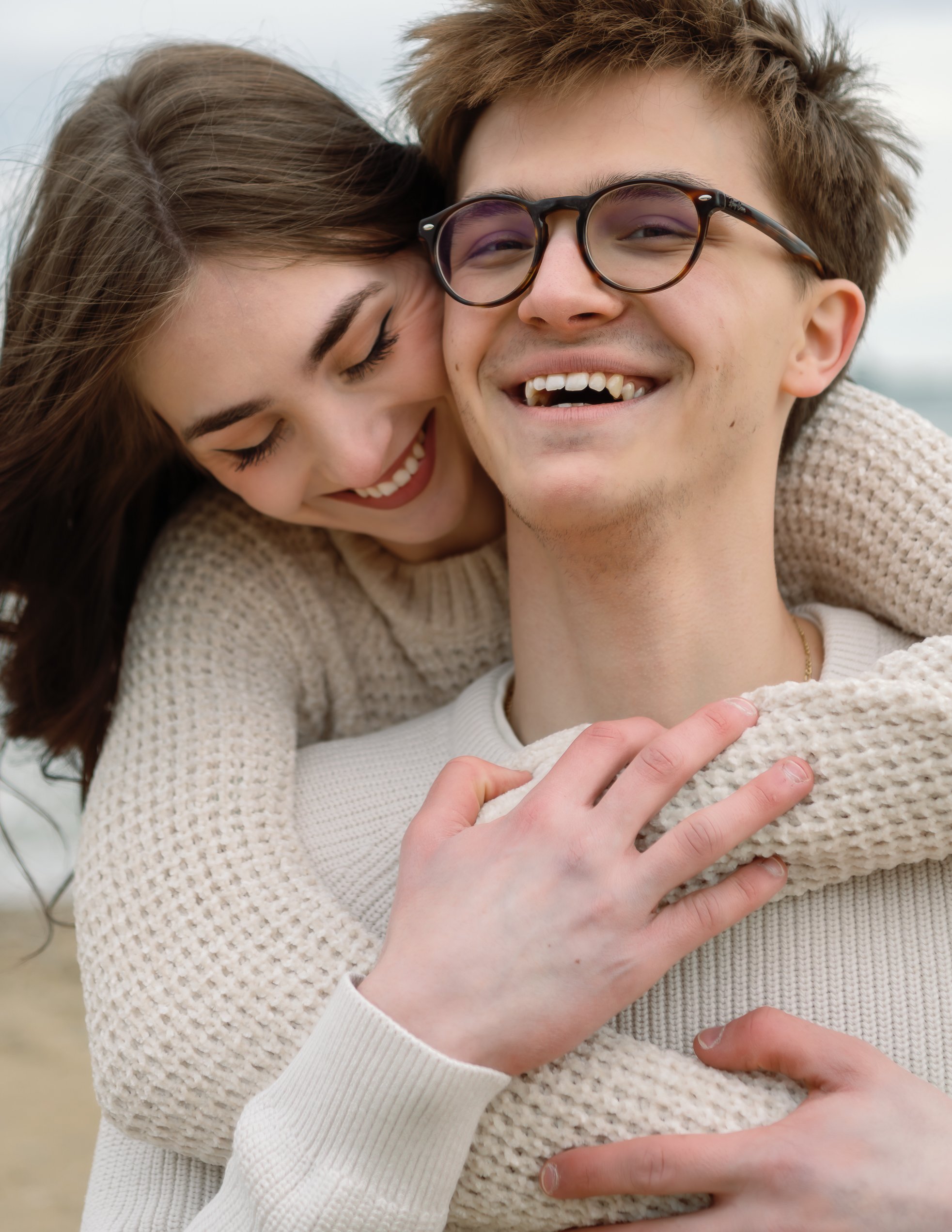 A young woman hugs a young man from behind, both smiling and appearing happy.