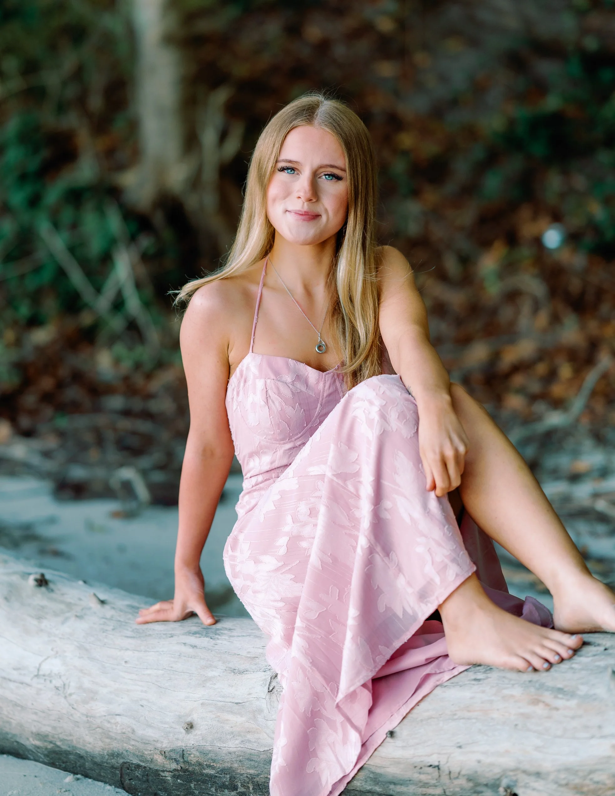 A woman in a pink dress sitting on a driftwood log by the water in a natural outdoor setting.