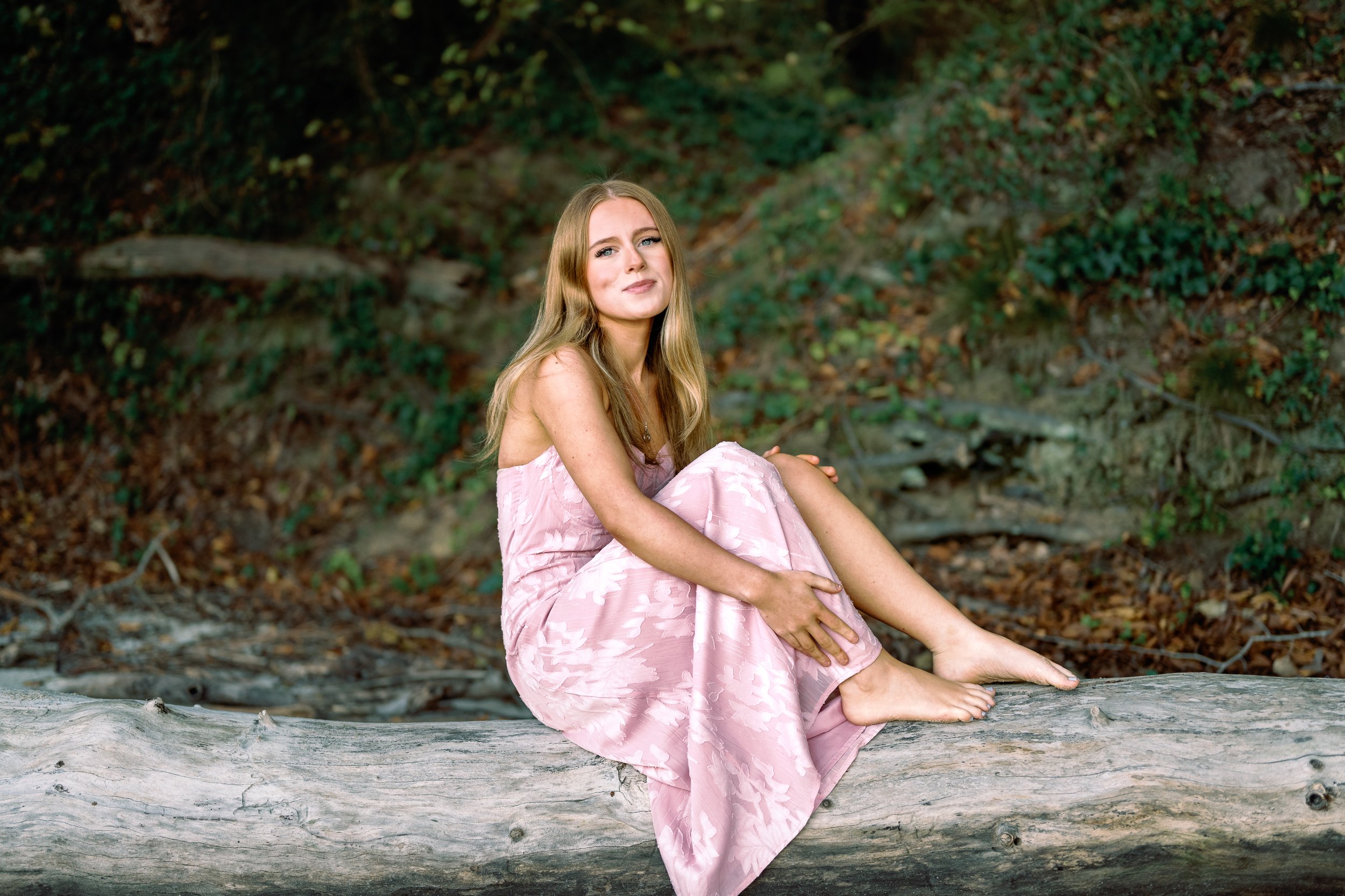 A young woman with long blonde hair wearing a pink dress is sitting on a fallen tree trunk in a forest, looking at the camera with a slight smile.