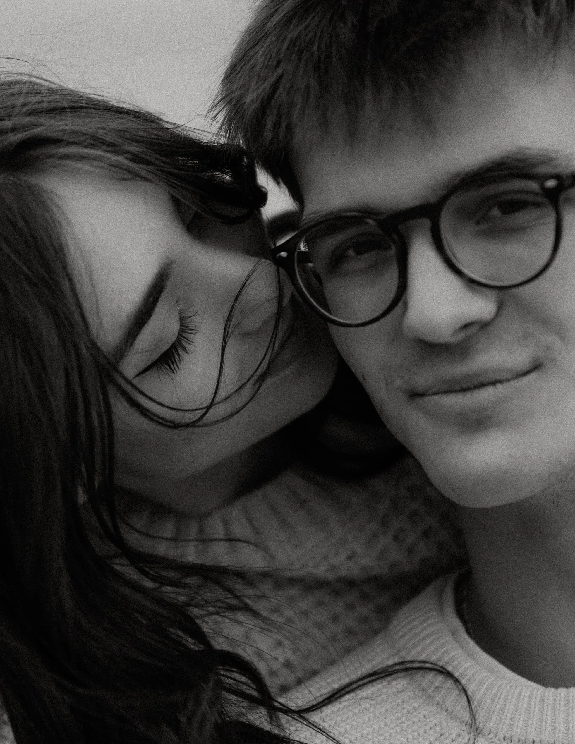 A black-and-white close-up photo of a young woman with wet hair kissing a young man with glasses and short hair on the cheek.