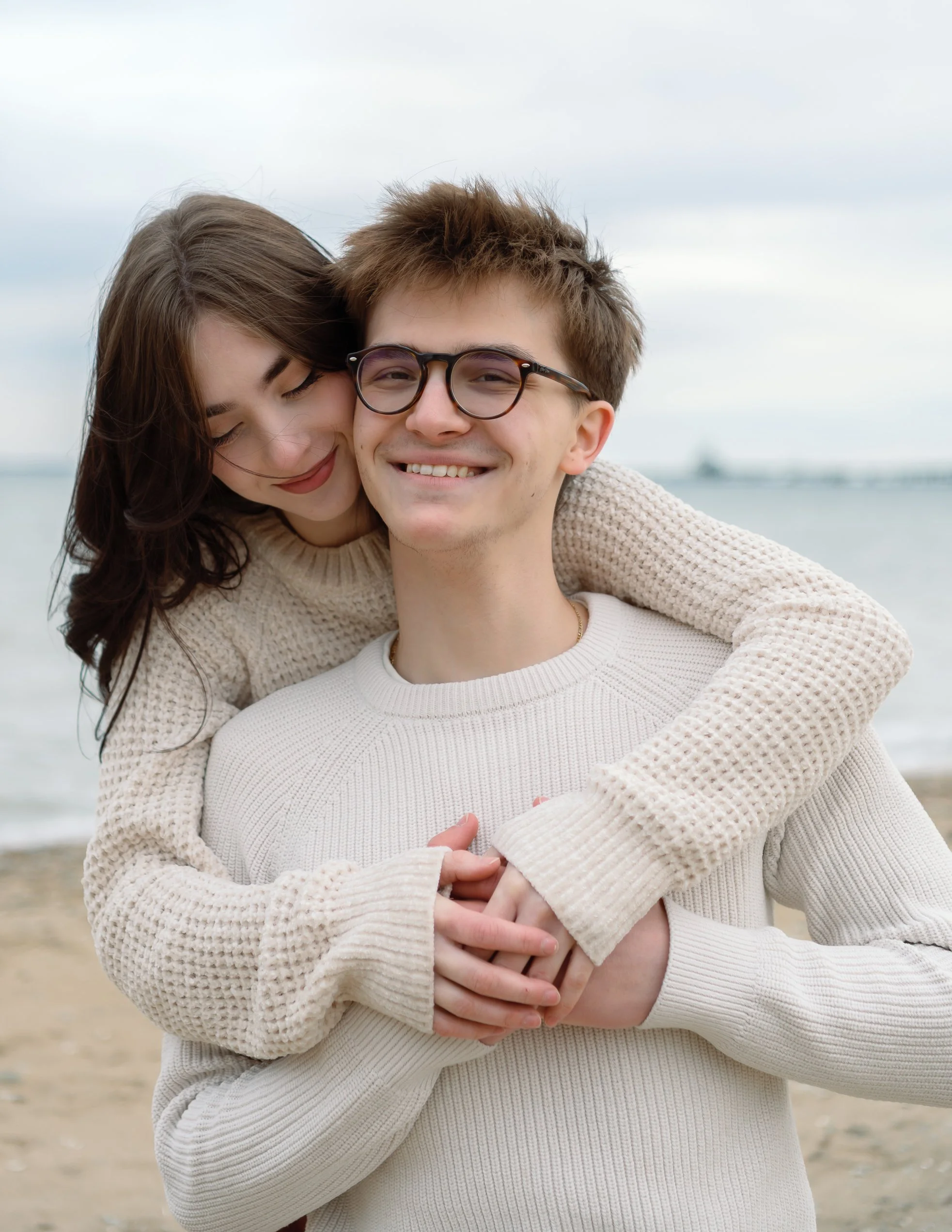 A happy young couple embraces on the beach, smiling at the camera, with a cloudy sky and water in the background.