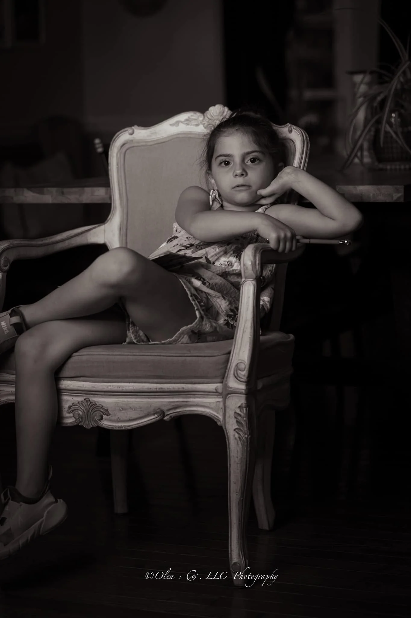 A young girl reclining on an ornate vintage armchair, resting her cheek on her hand, with a serious expression, in a dimly lit room.