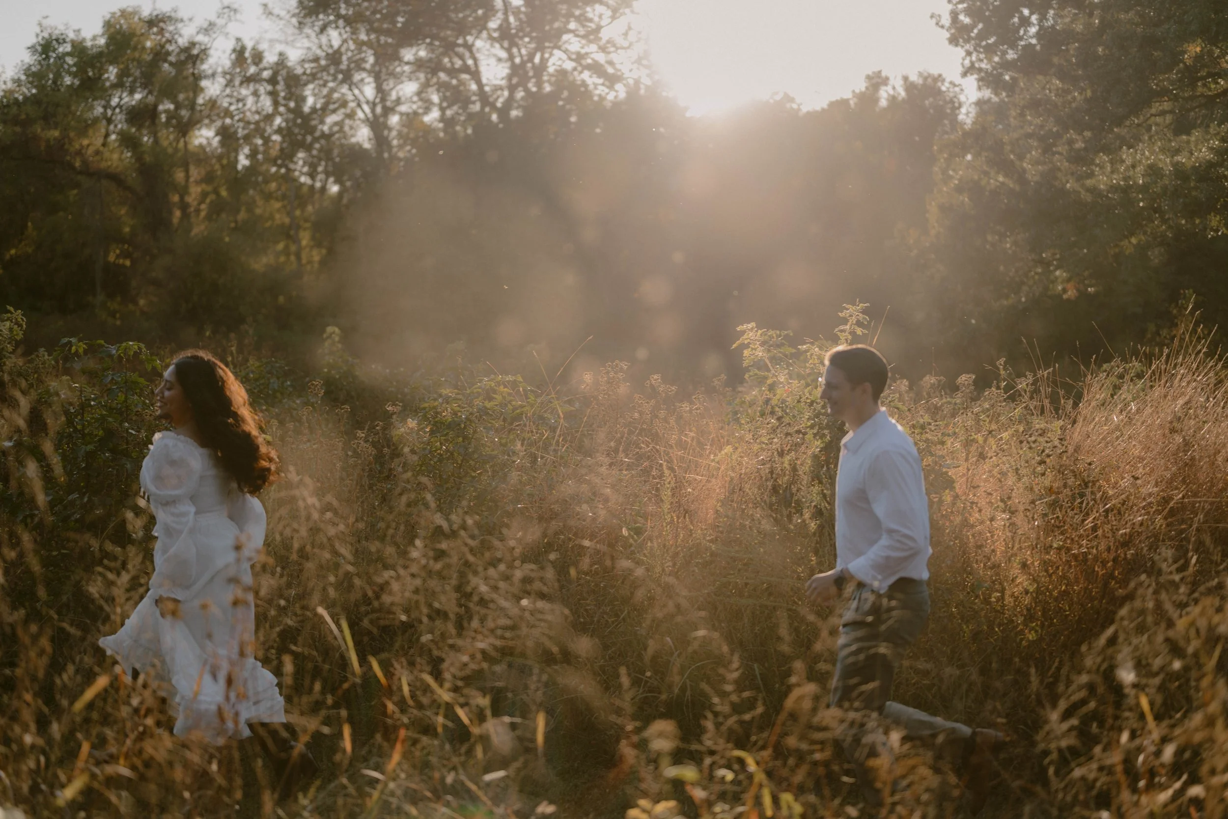 Couple running through tall grasses during a film-inspired engagement session at Chapman State Park in Indian Head, Maryland, backlit by warm autumn sunlight and surrounded by lush greenery.