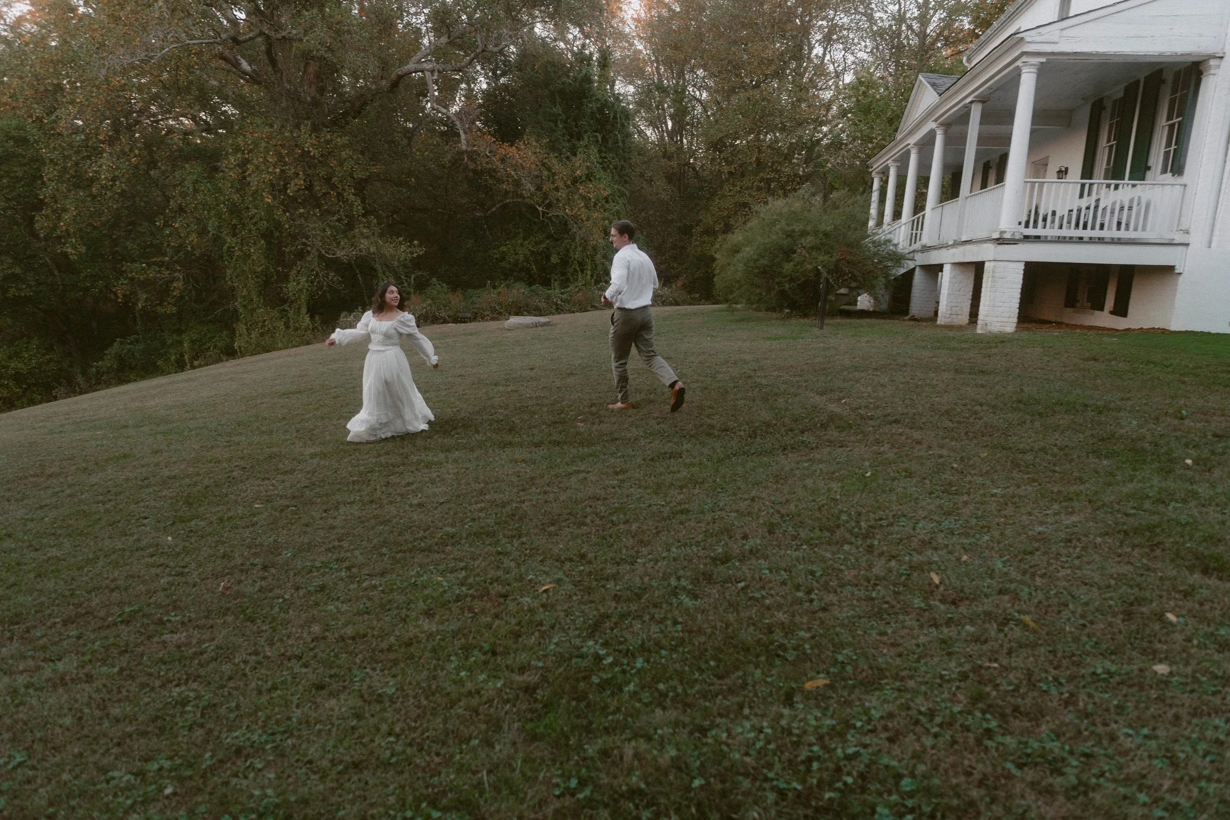 Couple running across a grassy lawn during a Pride & Prejudice–inspired engagement session at Chapman State Park in Indian Head, Maryland, with a historic white house in the background and autumn greenery surrounding them.