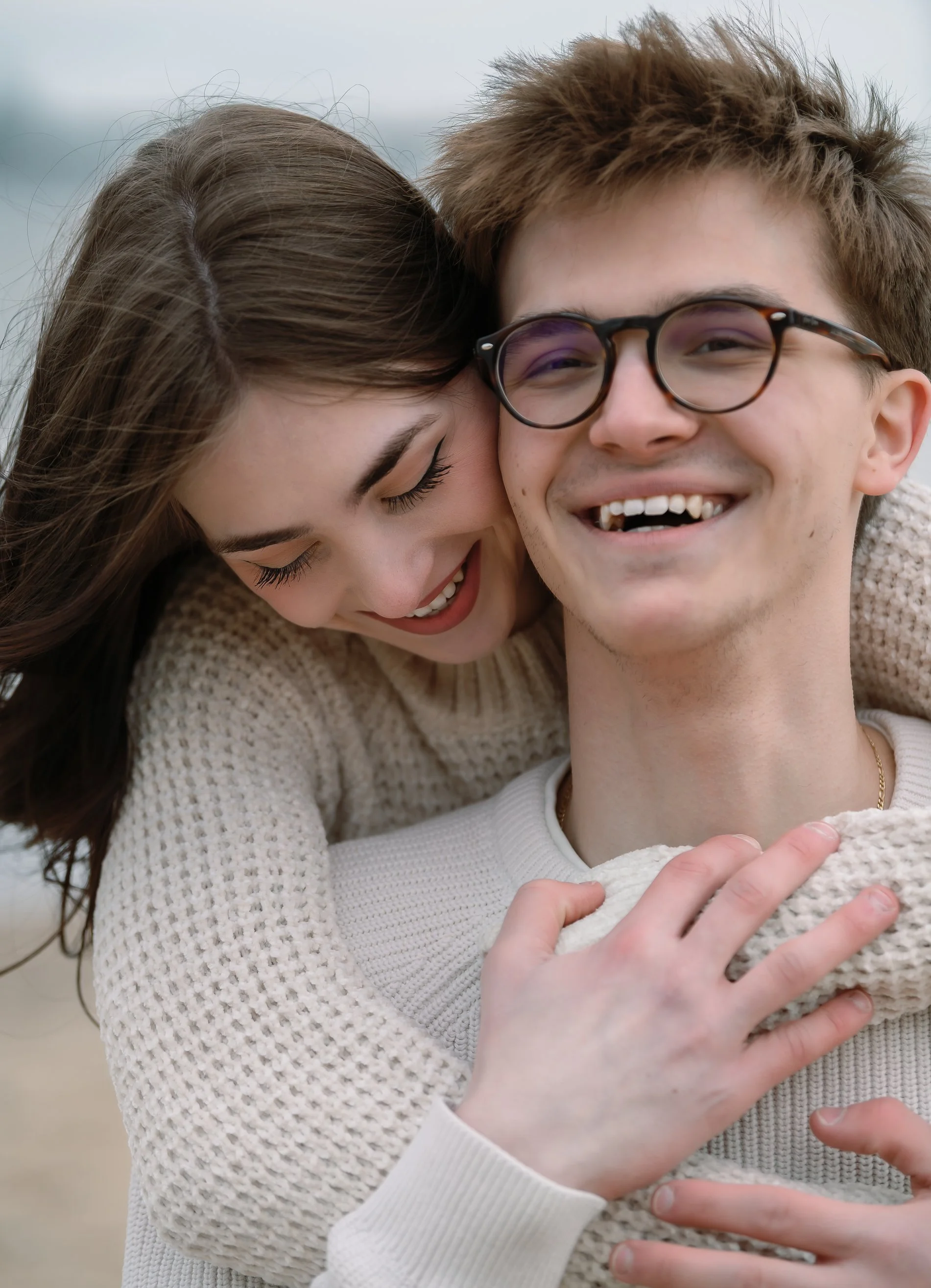 A young woman with brown hair is hugging a young man with glasses and short, light brown hair at the beach. They are smiling and appear happy.