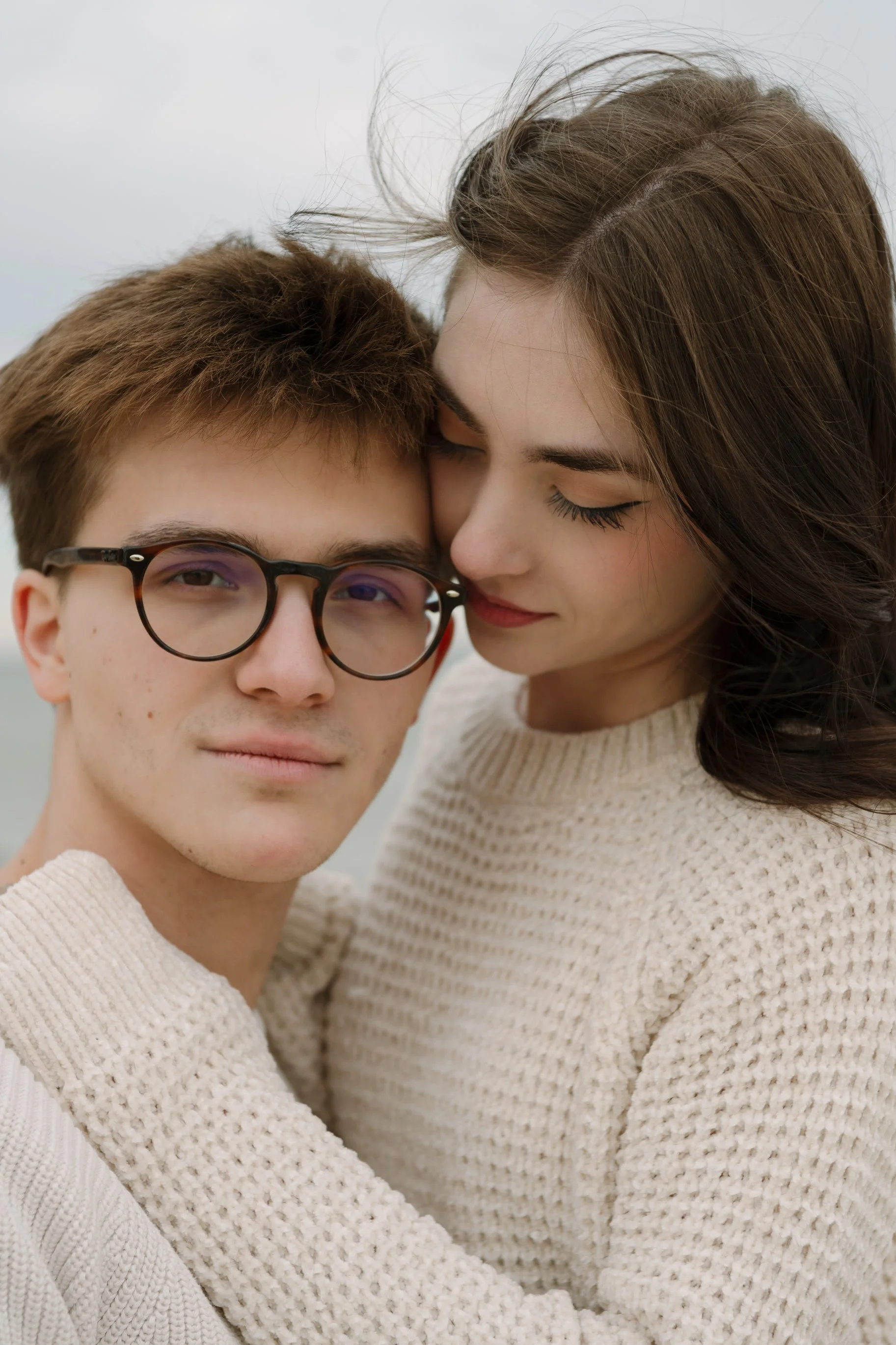 A young couple embraces outdoors, with the woman close to the man's face, her eyes closed and a gentle smile, while he looks at the camera wearing glasses.