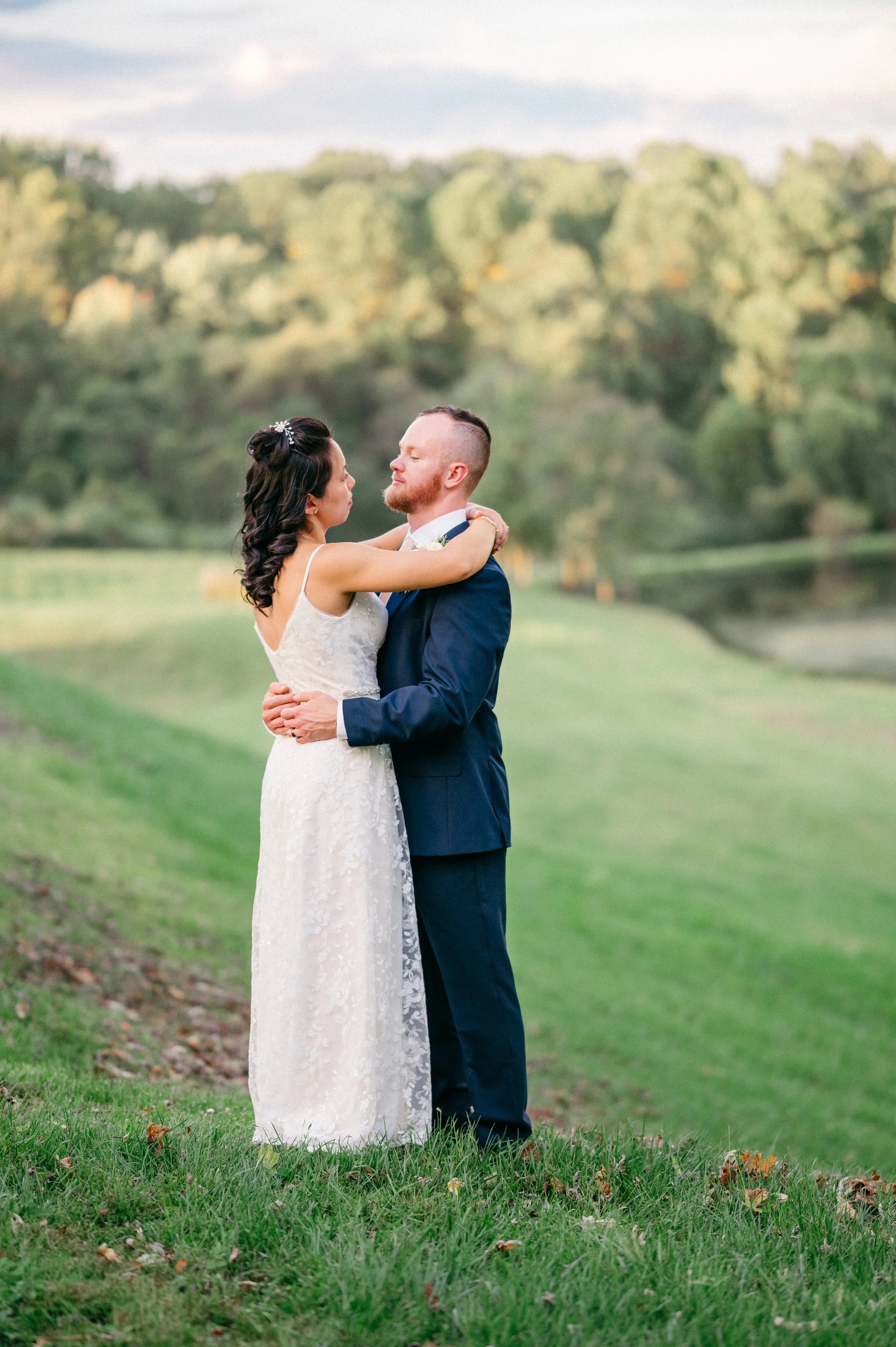 BRIDE AND GROOM HUG IN A FIELD WITH LAKE BEHIND THEM