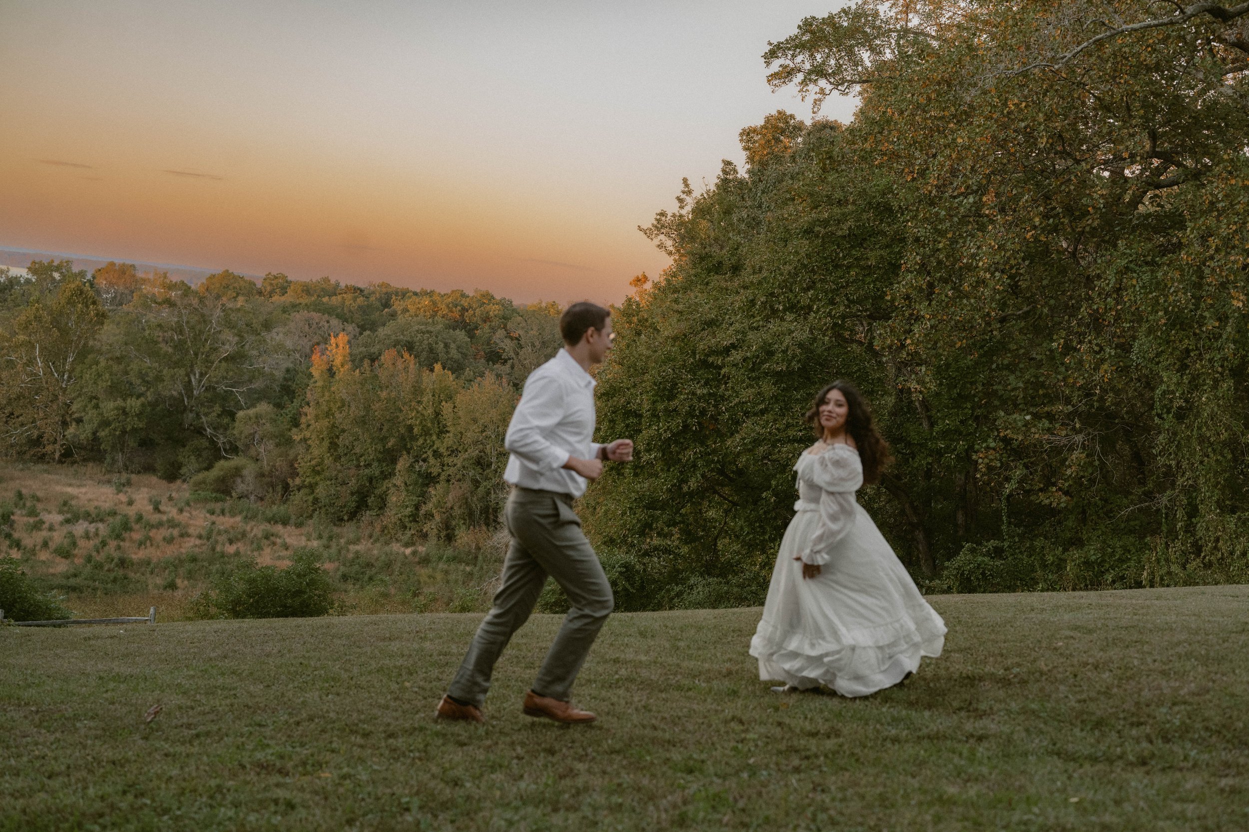 Couple running toward each other during a film-inspired engagement session at Chapman State Park in Indian Head, Maryland, with rolling hills, autumn trees, and a soft sunset sky in the background.