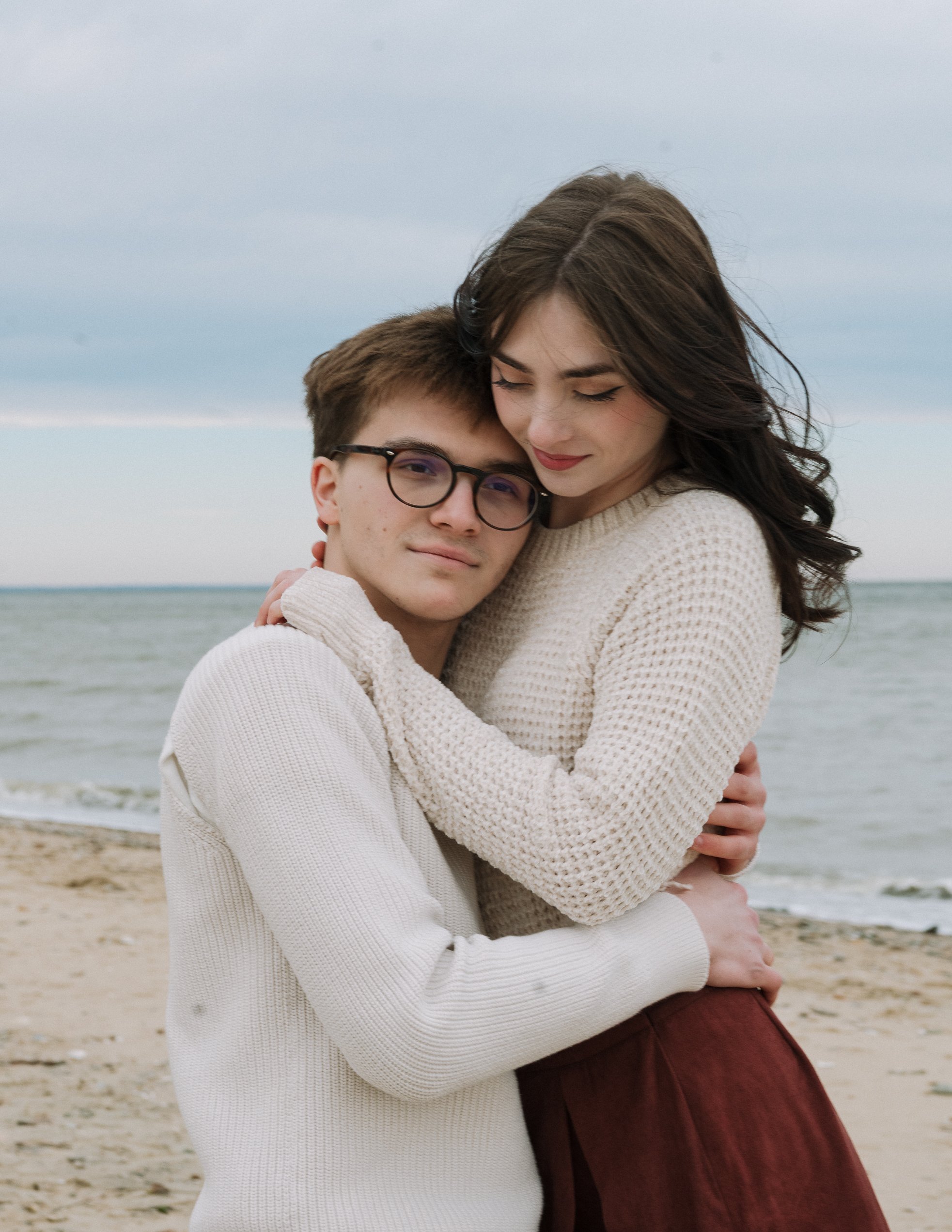 A young man and woman embrace on a beach, with the ocean and cloudy sky in the background.