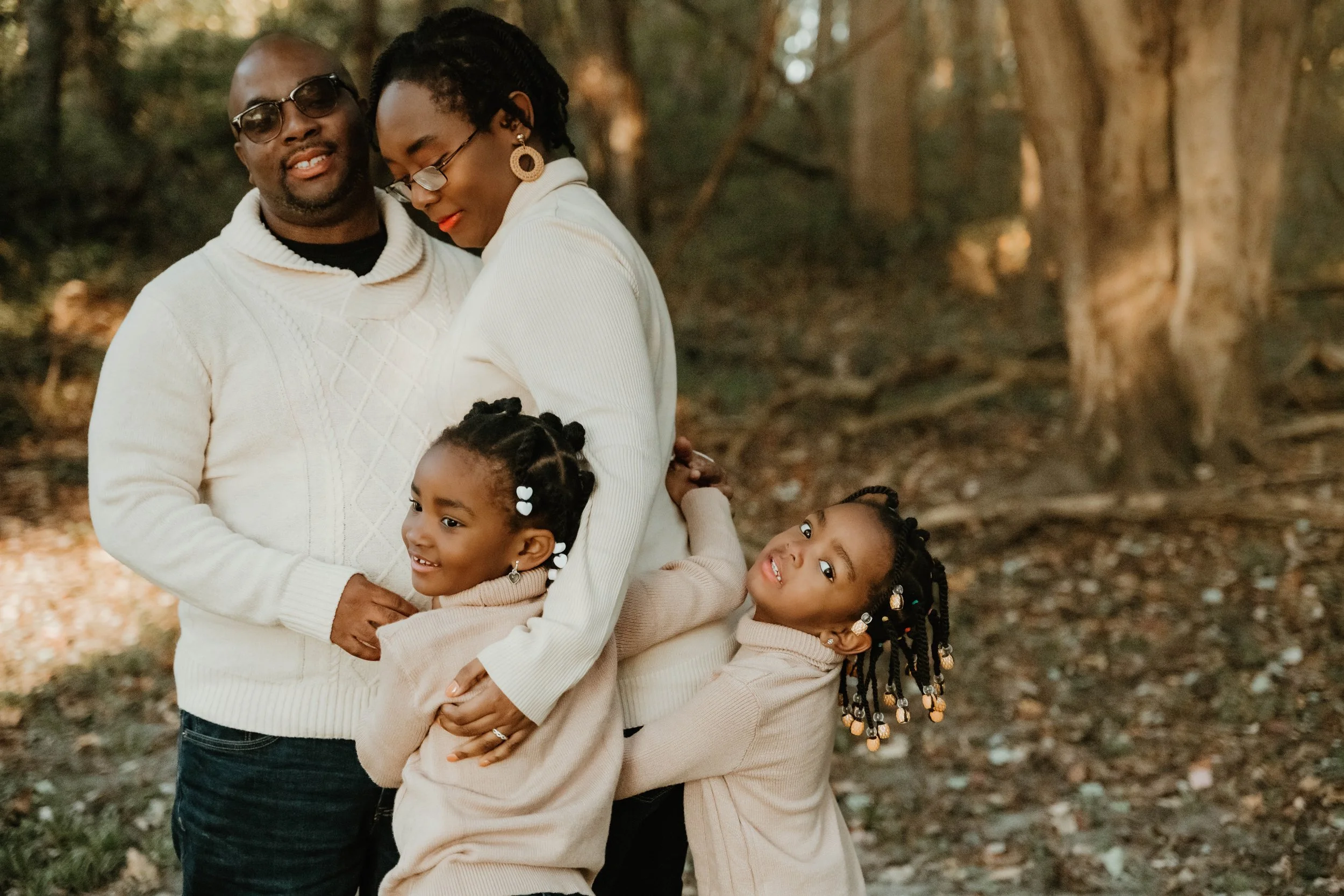 A family of four, including a father, mother, and two young daughters, hugging together outdoors in a wooded area during fall.
