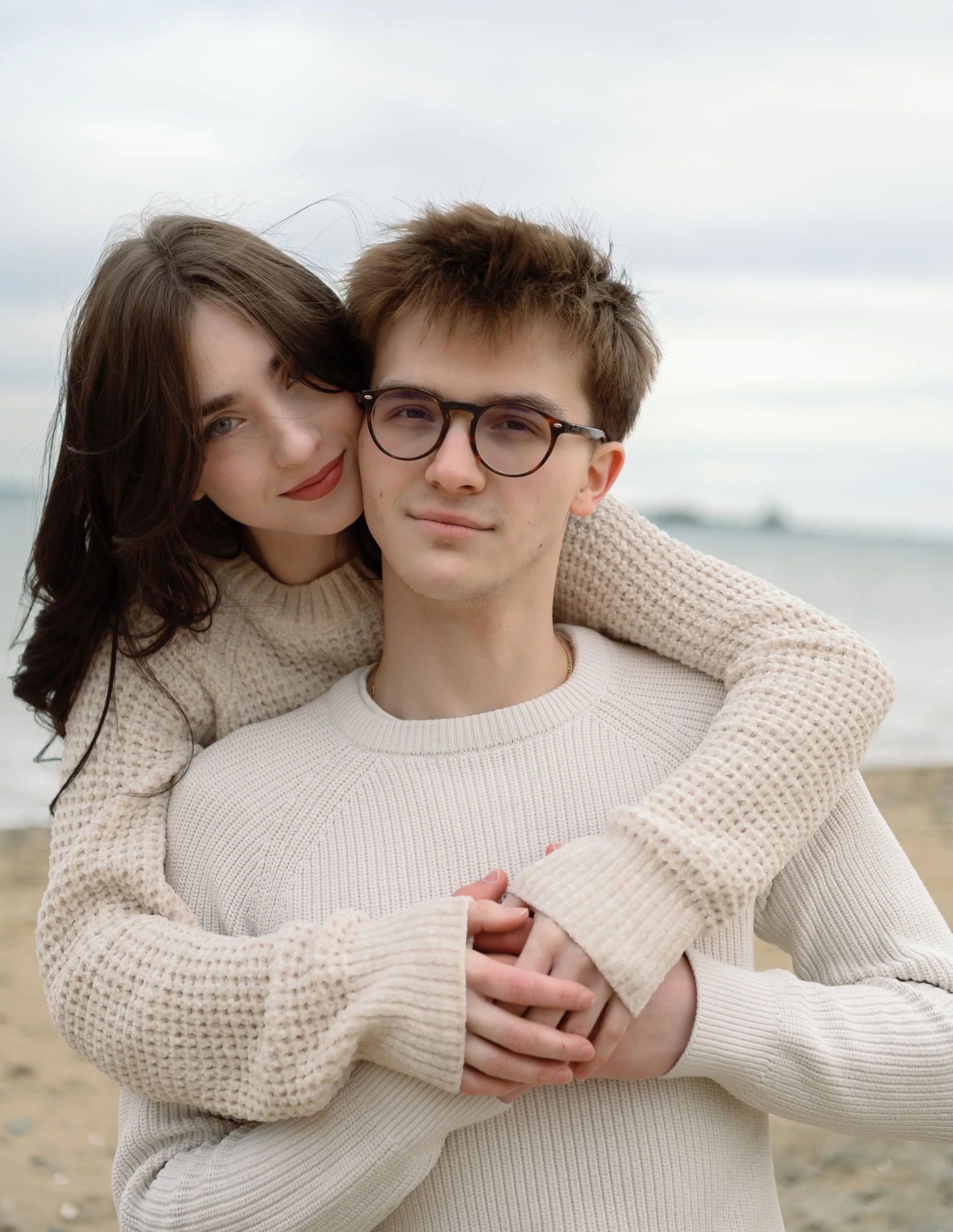 A young couple embracing at the beach, with the woman leaning on the man's shoulder, both smiling softly.