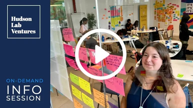 A group of people working on a collaborative project in a classroom or workshop setting. There are colorful sticky notes on the walls and glass, and a young girl smiling near the window. A video play button overlay is in the center.