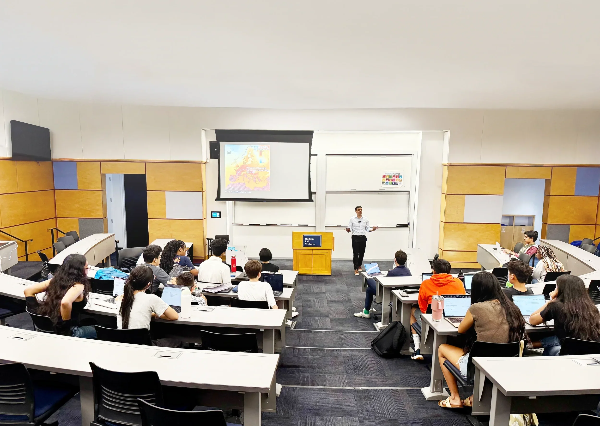 A university lecture hall with students listening to a male professor standing at the front, presenting a weather map on a screen, with students taking notes on laptops in a tiered classroom.