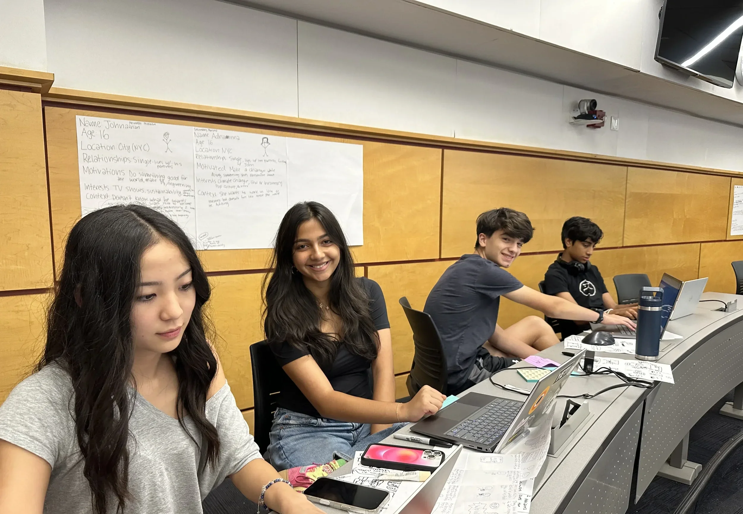 Four teenagers sitting at a computer desk in a classroom or conference room. They are working on laptops and have papers, phones, and a water bottle on the table. The is a wooden wall with posters behind them.