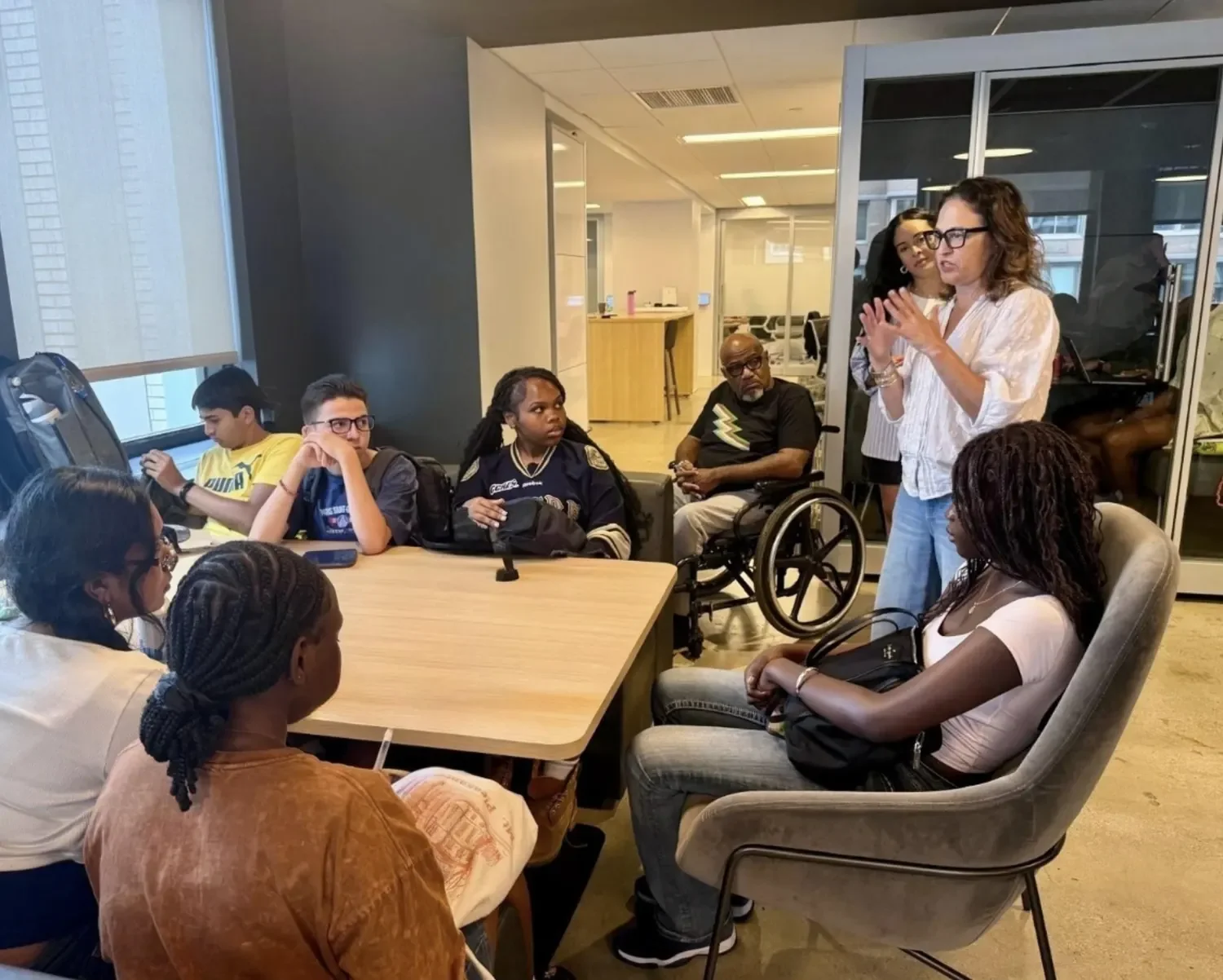 Group of diverse people in a meeting room listening to a woman who is speaking, with some seated at a table and others standing nearby, including a person in a wheelchair.