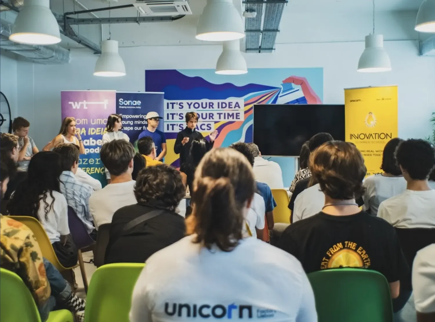 Group of young people presenting in front of an audience at a conference or seminar with colorful banners and a large screen behind them.
