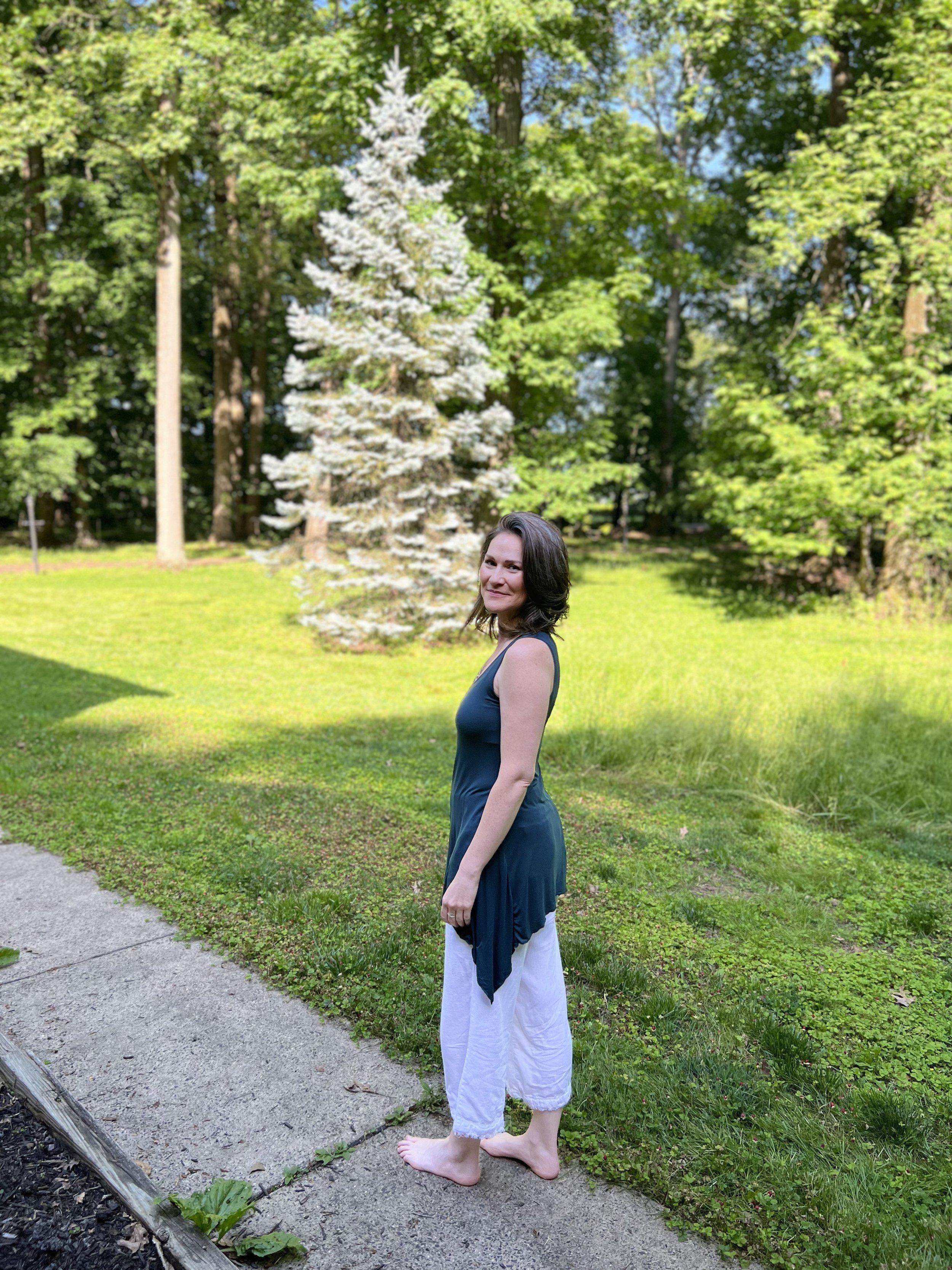 Woman standing on a concrete path in a garden, wearing a teal top and white pants, with a lush, green background and a small fir tree.
