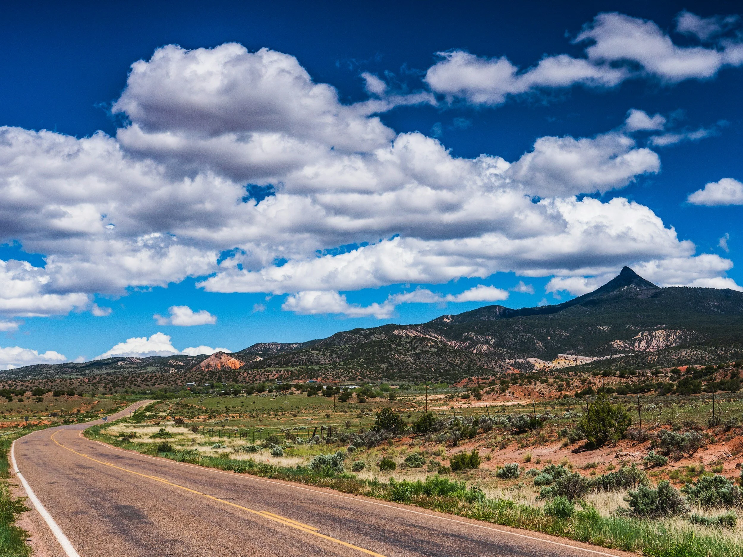 Scenic road leading towards mountains under a blue sky with fluffy clouds.