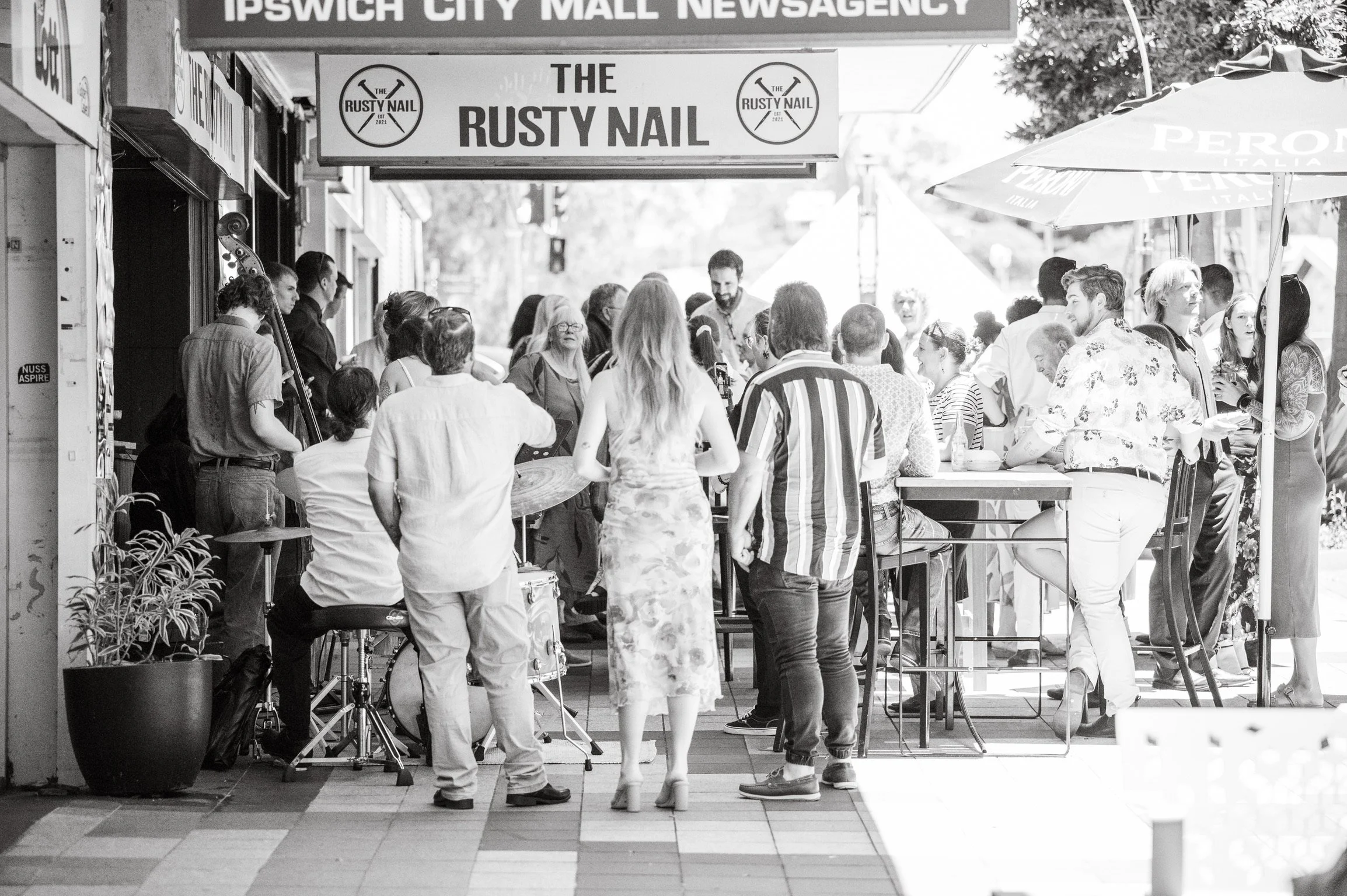 A group of people gathered outside a cafe or bar under an awning, with some standing and some sitting at tables, engaging in conversation. There are signs and umbrellas visible, with a sign reading 'The Rusty Nail' and another sign mentioning Ipswich City Mall Newsagency. The scene appears lively and social on a bright day.