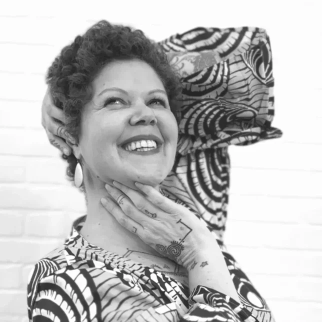 A smiling woman with curly hair, wearing earrings, with her hand resting on her neck, and a zebra-patterned fabric behind her.