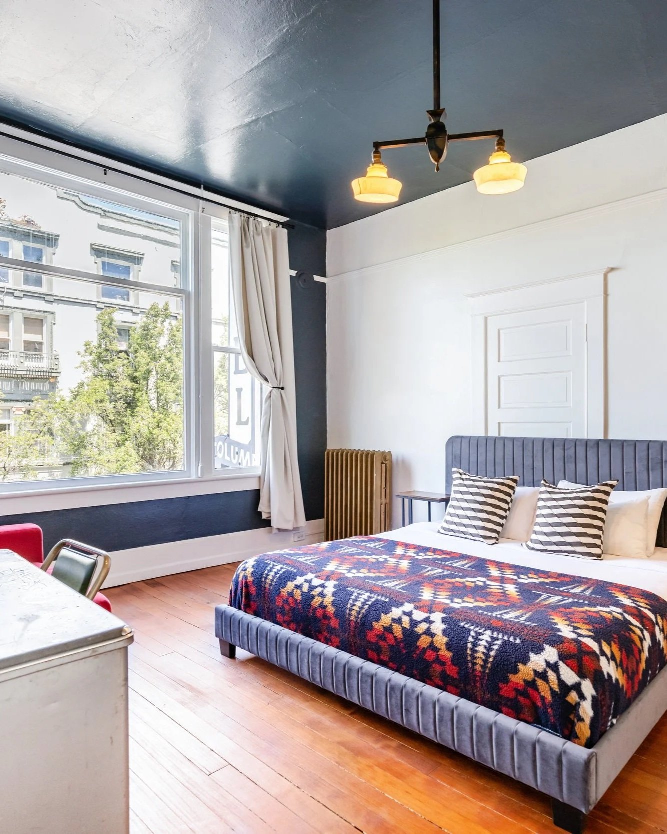 A room with wood floors and a large window facing the main street. The bed is large with white sheets and a comfortable heavy blanket. A desk sits to the side.