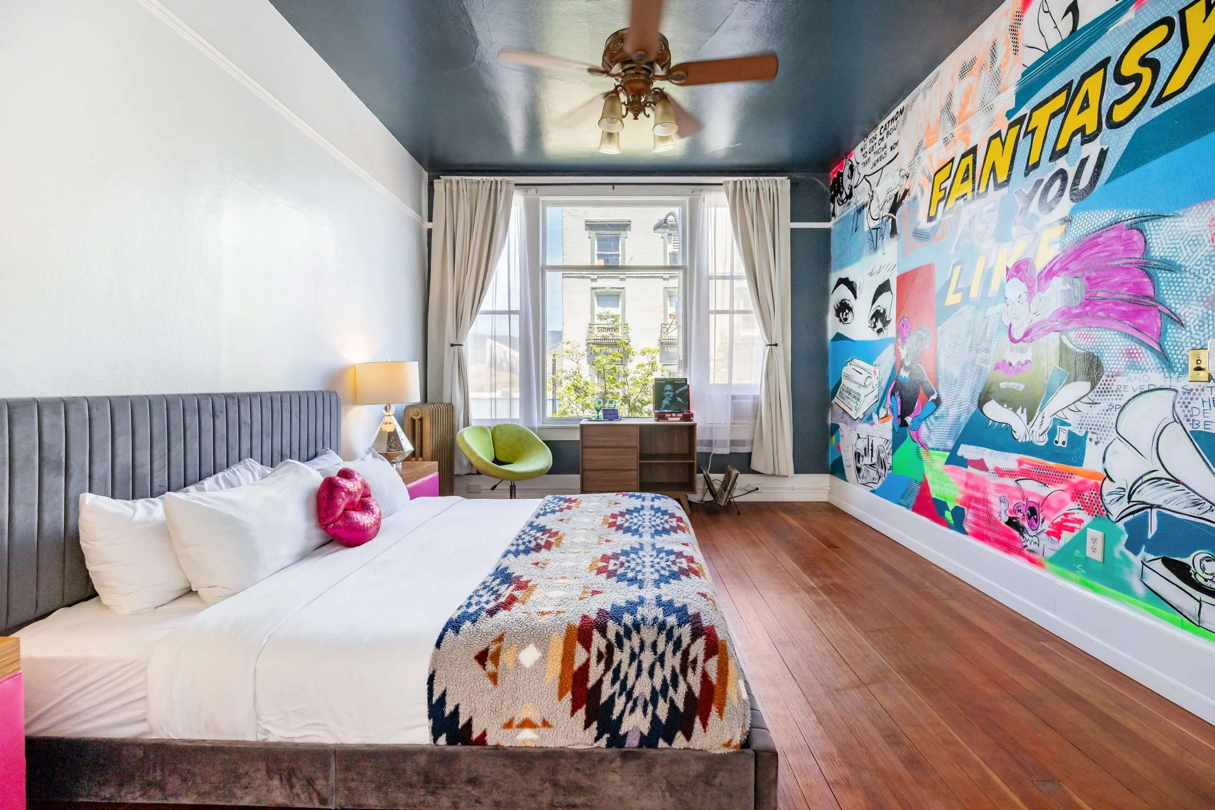A bedroom with a bed featuring a gray upholstered headboard, white bedding, and pink lips-shaped pillows. A colorful quilt is at the foot of the bed. Next to the bed is a wooden nightstand with a lamp. The room has a large window with white curtains, allowing natural light in, and there's a lime green chair and a small wooden cabinet. The right side wall has vibrant, comic-style artwork with various illustrations and text