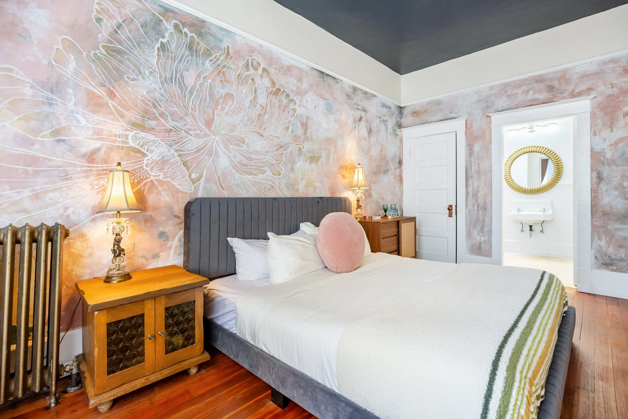 A bedroom with a bed, nightstands, a chair near the window, wooden flooring, and a wall with floral mural.