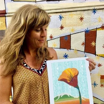 Woman holding a colorful painting of a flower with an orange bloom in front of a city mural.