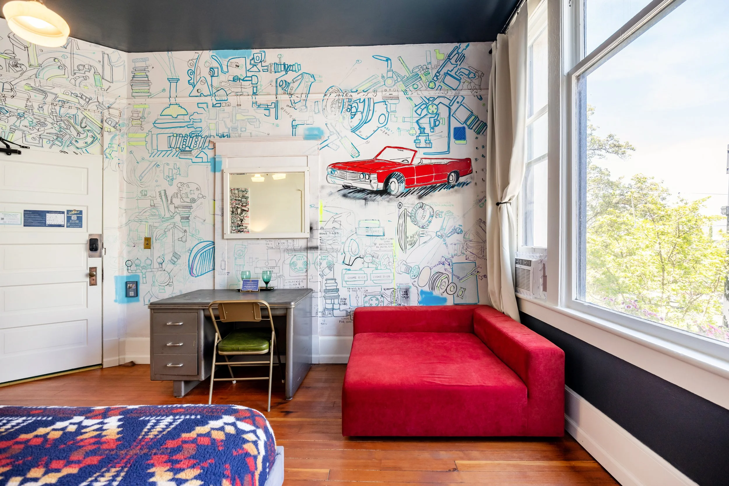 Room interior with a wall decorated with automotive sketches and a red vintage car mural, a desk with a green chair, a red couch, and large windows letting in natural light.