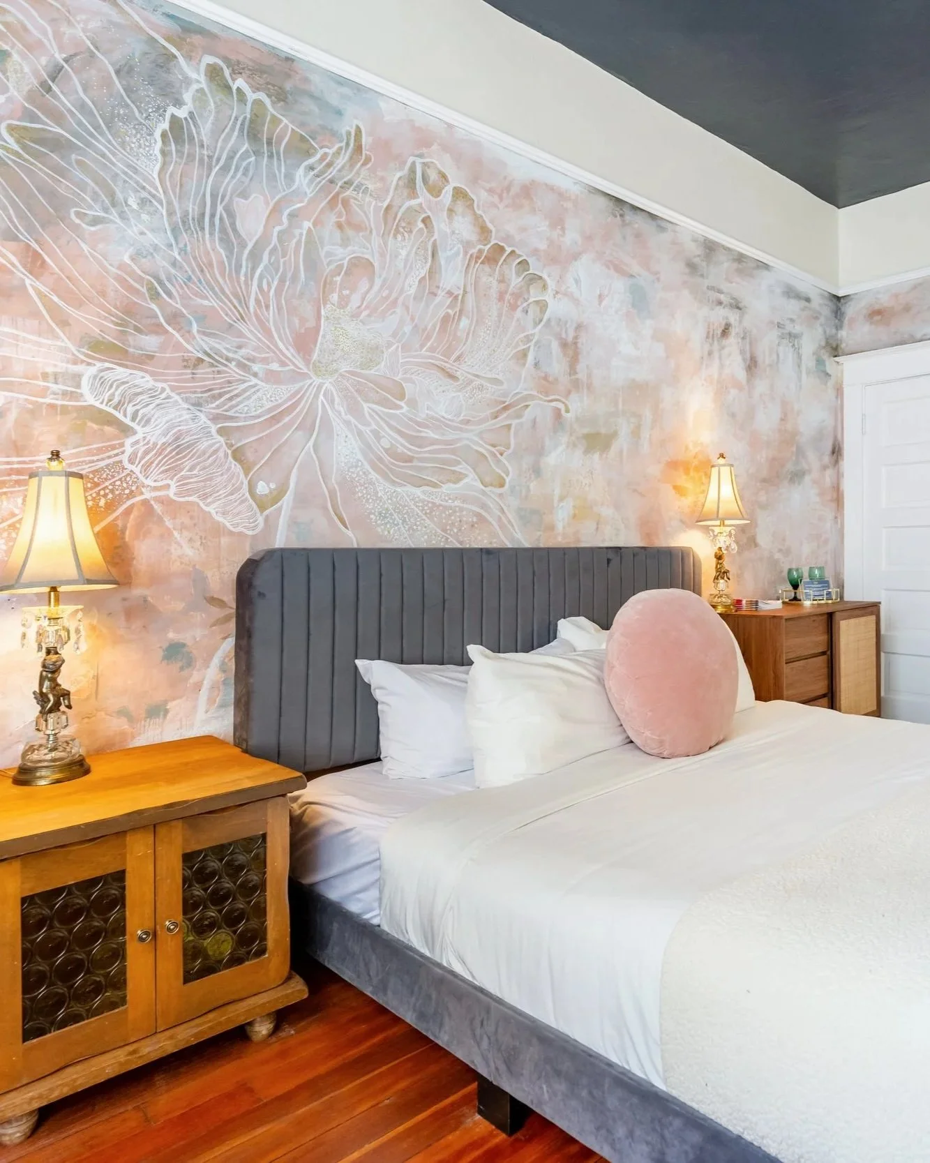 A cozy bed with white sheets and a white heavy blanket and a pink throw pillow. Two dressers sit beside it with lit decorative lamps. The wall behind the bed is painted with a pale pink floral mural. The floor is wooden.