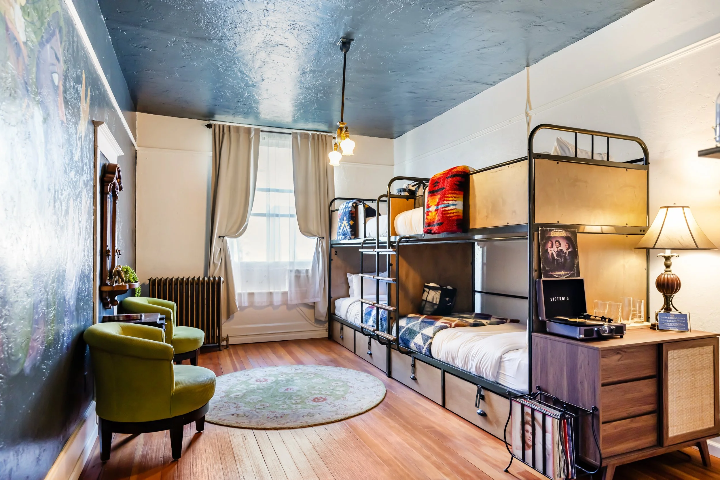 A cozy room with wooden flooring, containing two sets of bunk beds, a yellow armchair, a small oval rug, a window with curtains, an antique mirror, a radiator, and a wooden dresser with bottles and cups.
