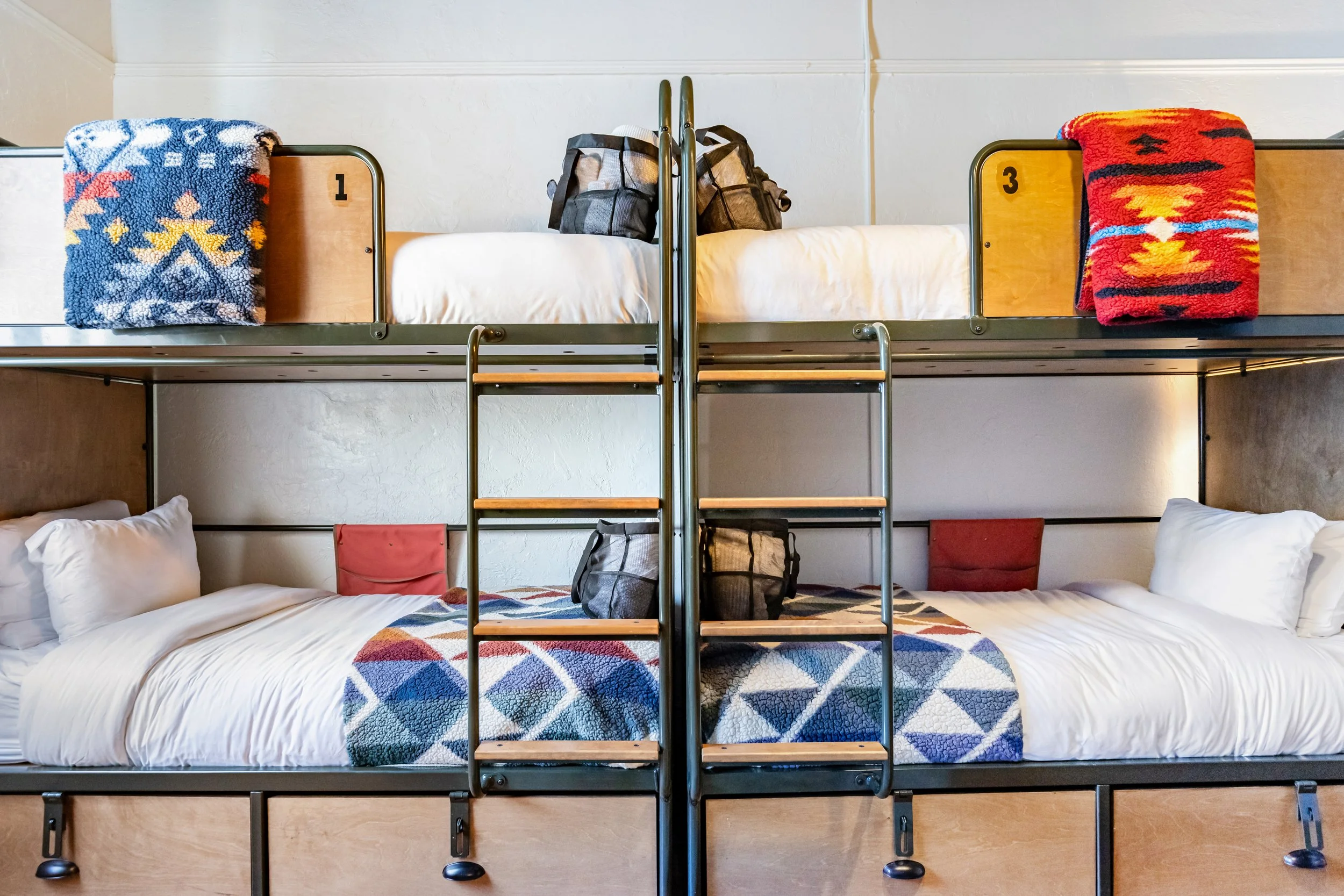 Set of two comfortable adult size bunk beds pushed up together against a wall with white sheets and cozy heavy blankets. They also have bags with towels and robes.