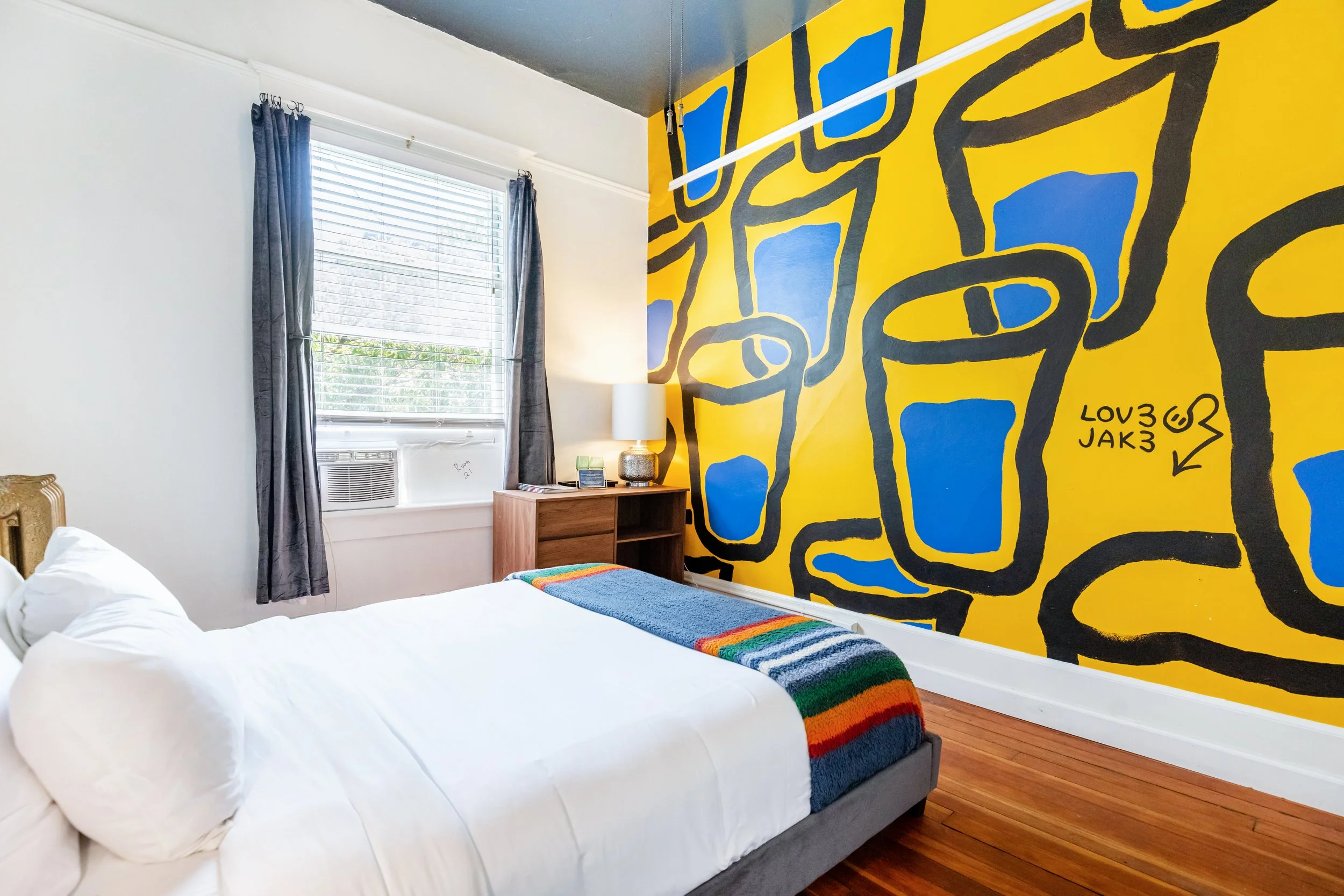 A bedroom with a white bed, colorful rainbow throw blanket, and a vibrant yellow wall with bold black and blue abstract graffiti art. There is a window with gray curtains, a small wooden nightstand with a lamp, and hardwood flooring.