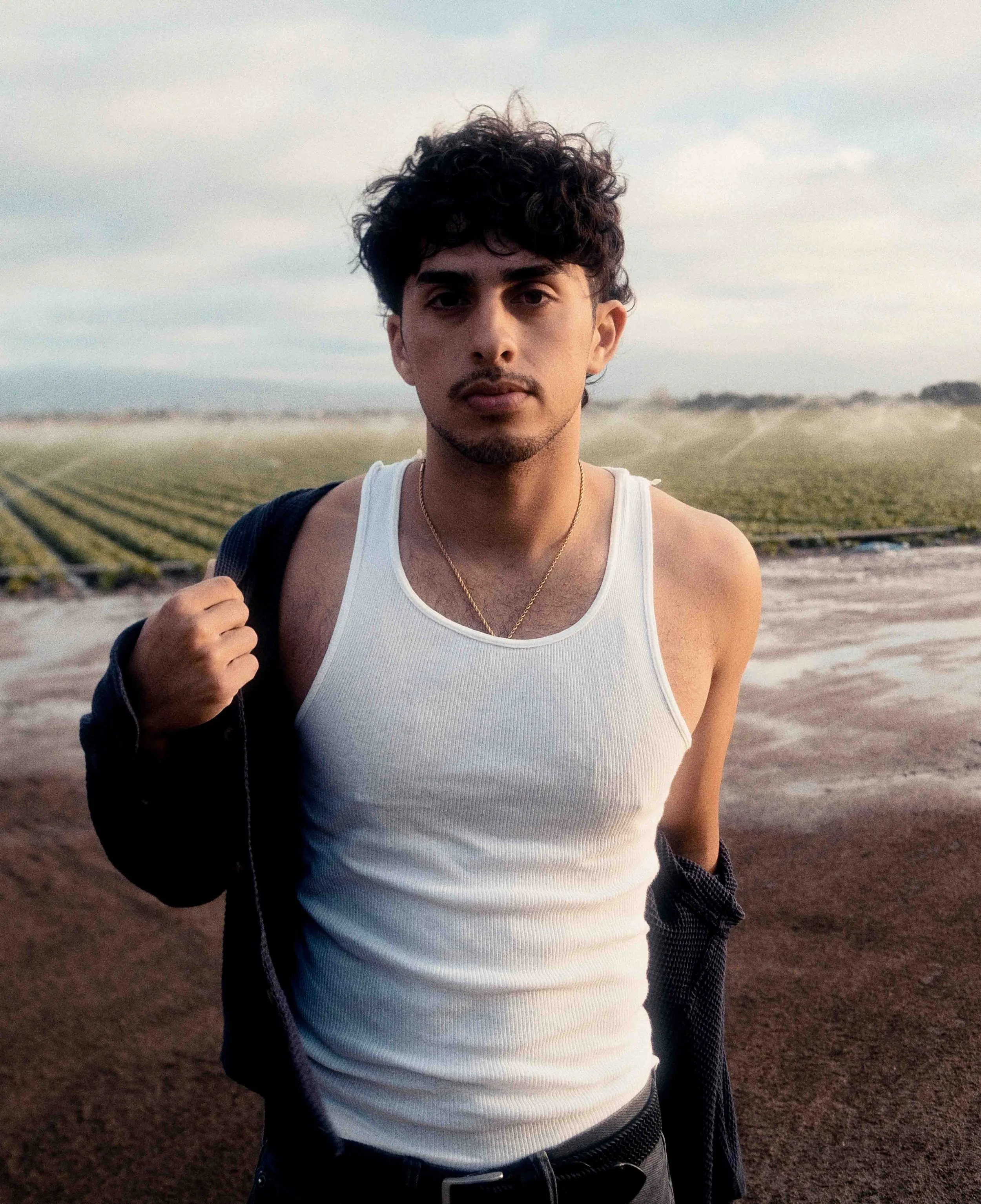 A young man with curly dark hair and a mustache, wearing a white tank top and a necklace, standing outdoors in a field with rolling hills and cloudy sky in the background.