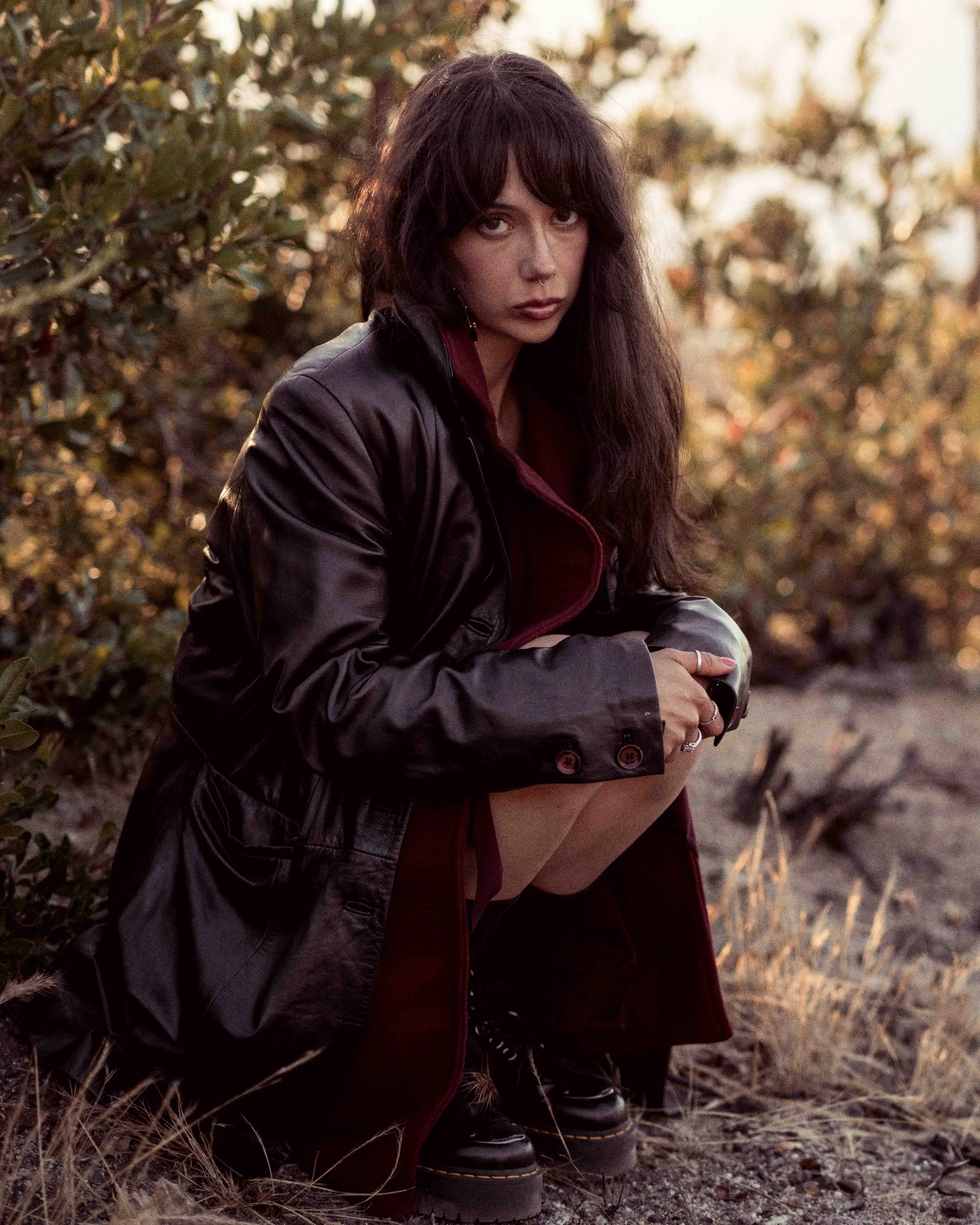 A young woman with dark, wavy hair and a septum piercing sitting outdoors near bushes, with a blurred natural background during sunset, wearing a dark leather jacket over a maroon shirt, slightly leaning forward with her arms resting on her knees.