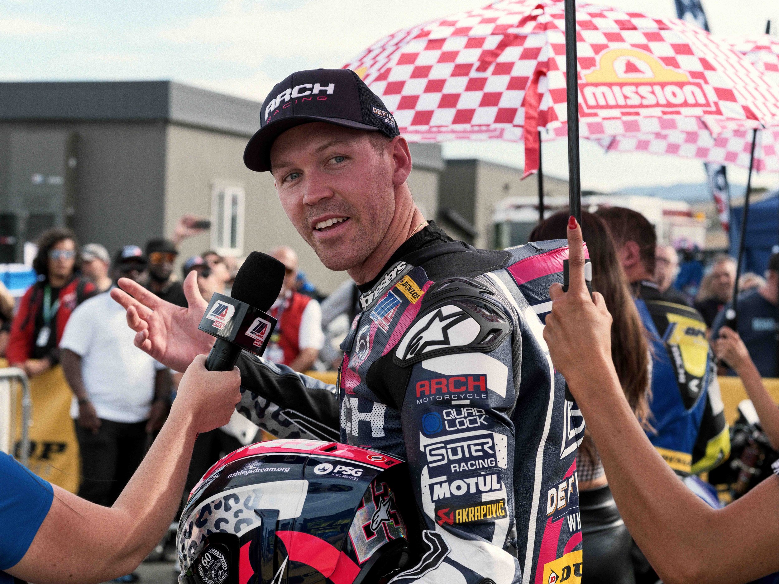 A race car driver wearing racing gear is being interviewed after a race, with a crowd of spectators and team members behind him at the race track. He is holding a helmet and gesturing with one hand, while another person holds a microphone towards him