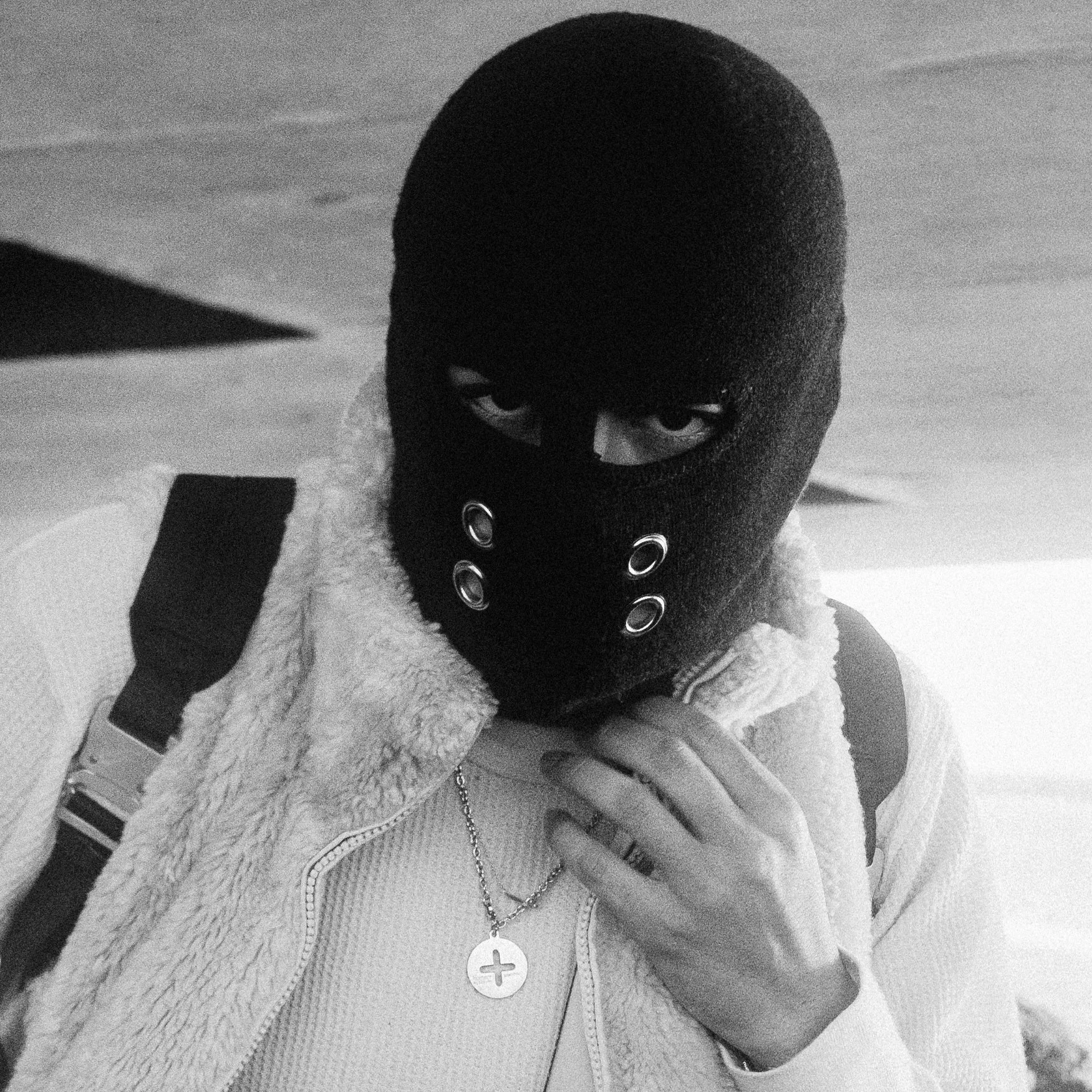 Person wearing a black ski mask, white fur-lined jacket, layered necklaces, and a backpack, taking a selfie indoors in black and white.