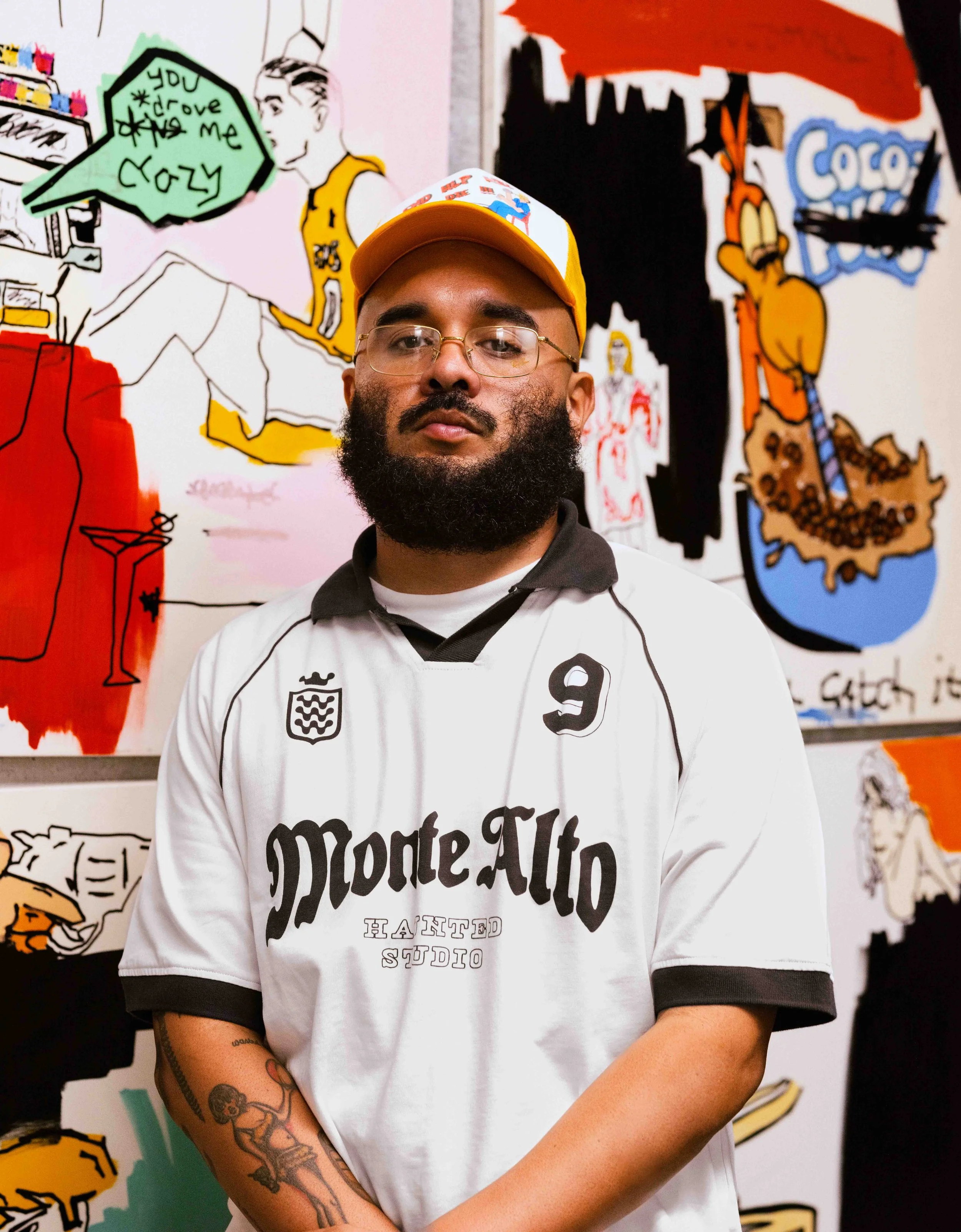 A man with glasses and a beard wearing a white Monte Alto T-shirt and a yellow cap, standing in front of colorful cartoon-style murals of characters and speech bubbles.