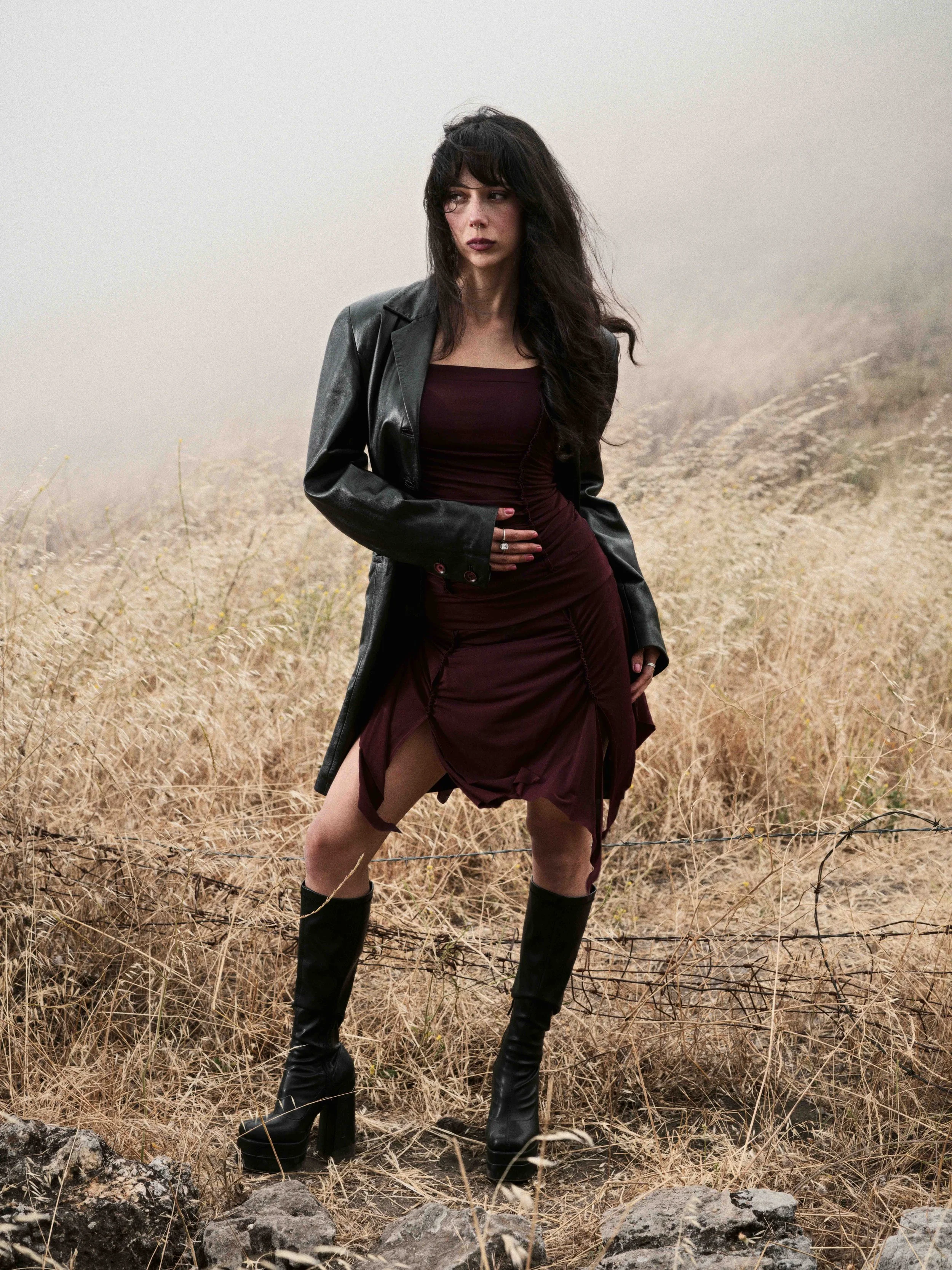A woman with long dark hair, wearing a black leather jacket over a burgundy dress, black thigh-high boots, and rings, standing in a foggy, dry grassy field with rocks and barbed wire.